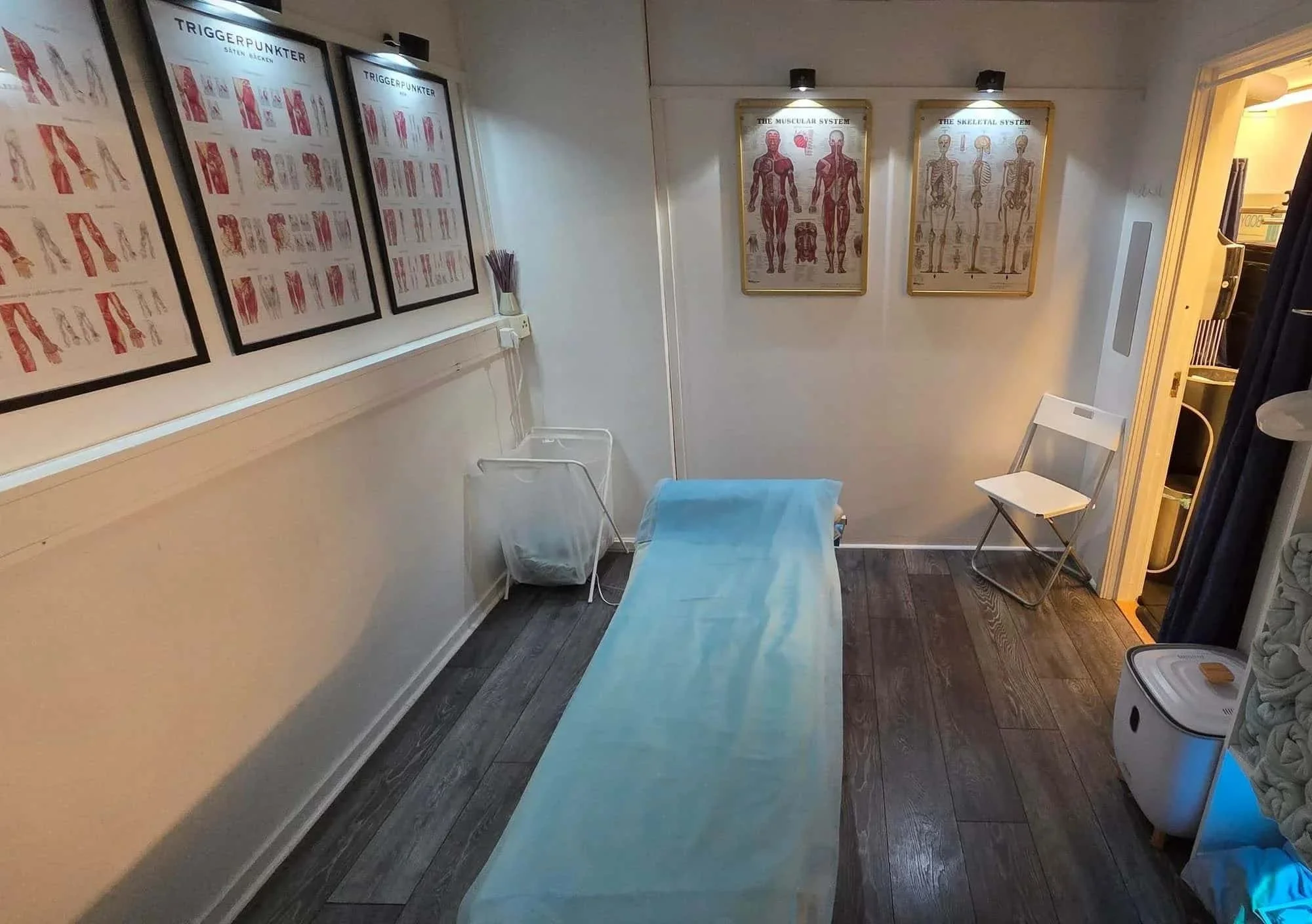 Behandlingsrum med en massagebänk täckt med en blå lakan, tre stolar, diplom från universitet och kursbeskrivningar på väggar, en hylla med handdukar och anatomi- samt triggerpunktsplanscher på väggarna.