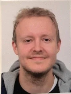 Porträtt av en man med ljus hår och lätt skägg, bär en grå hoodie.