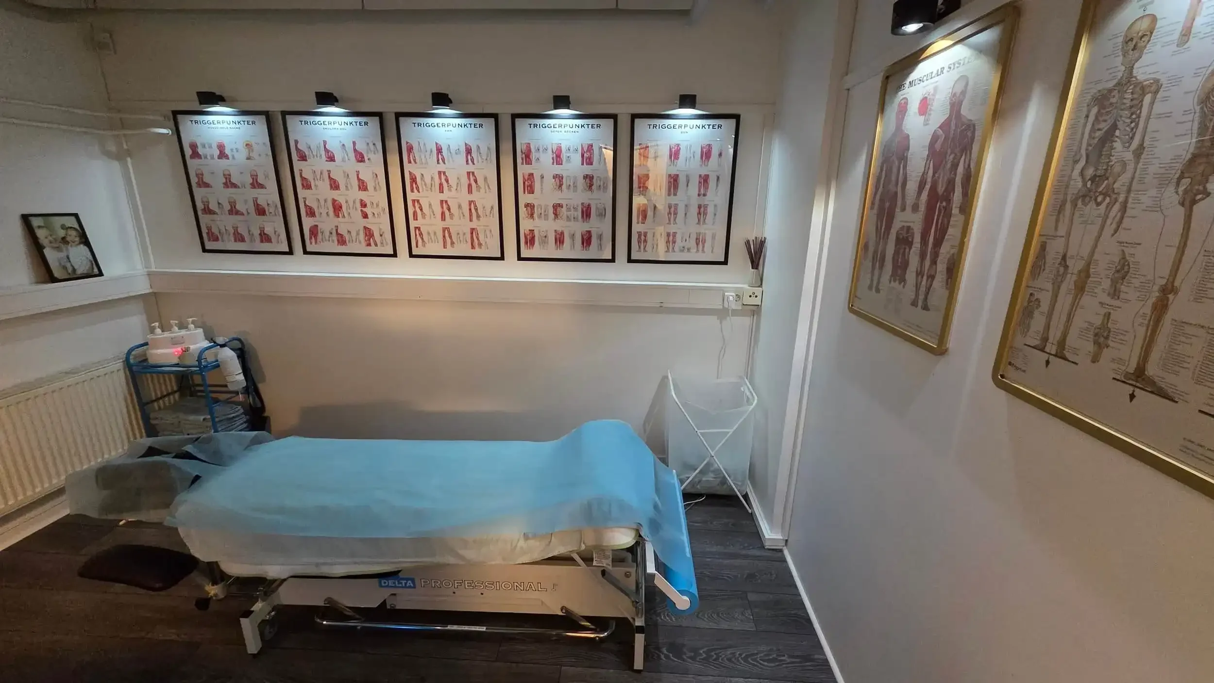 Massagebänk i ett massagerum med bildark, massageutrustning och muskeldiagram på väggarna.