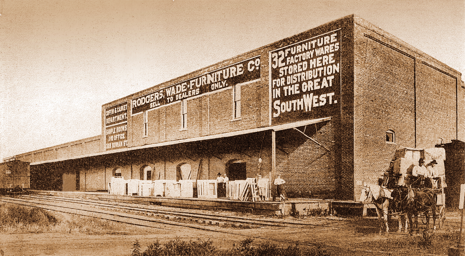 Historical black-and-white photo of a large brick building with signs advertising furniture and factory wares, and people and horses standing at the entrance.
