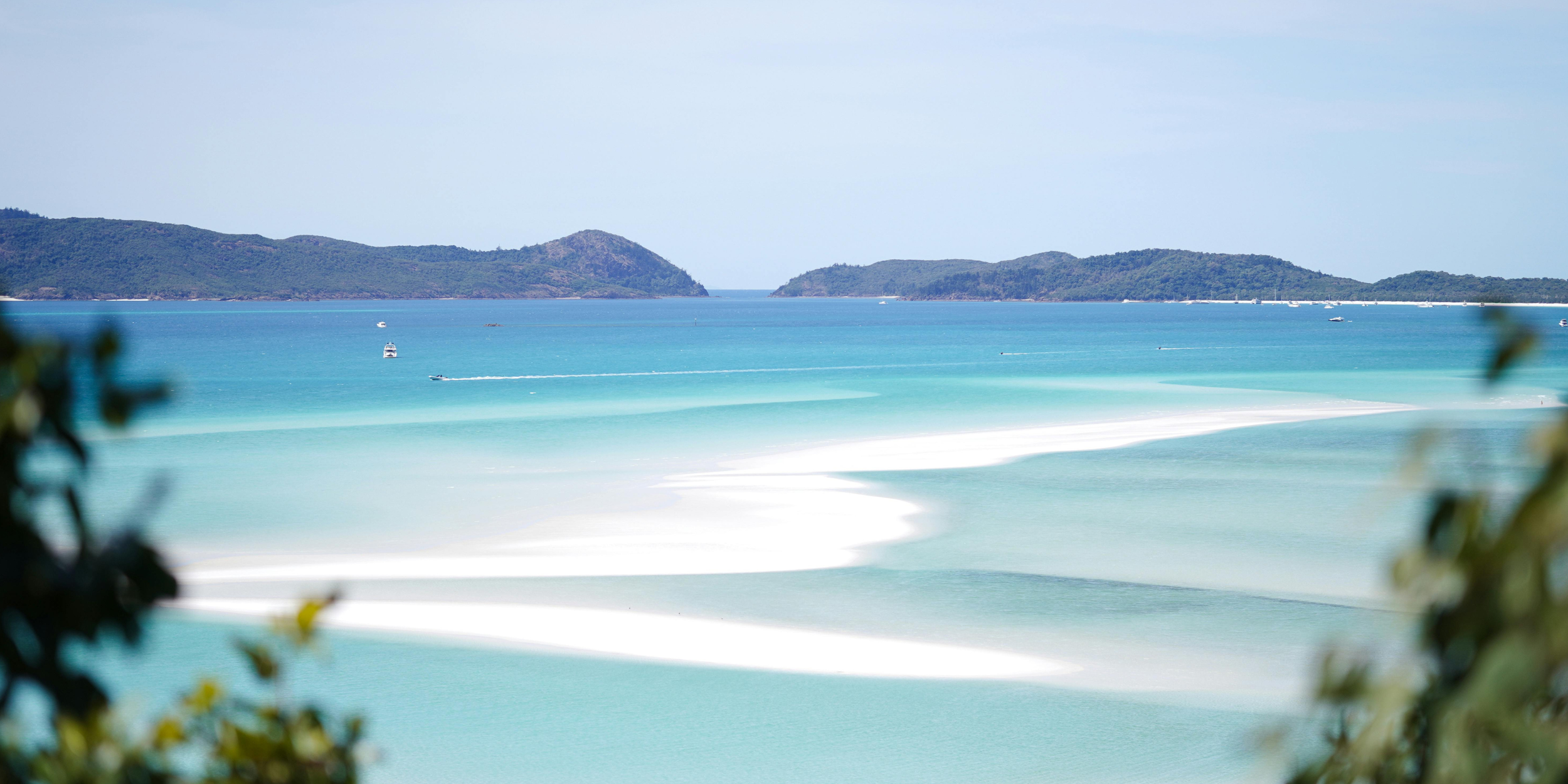 Scenic view of a tropical beach with white sand, turquoise water, and green islands in the distance, framed by blurred leaves in the foreground.