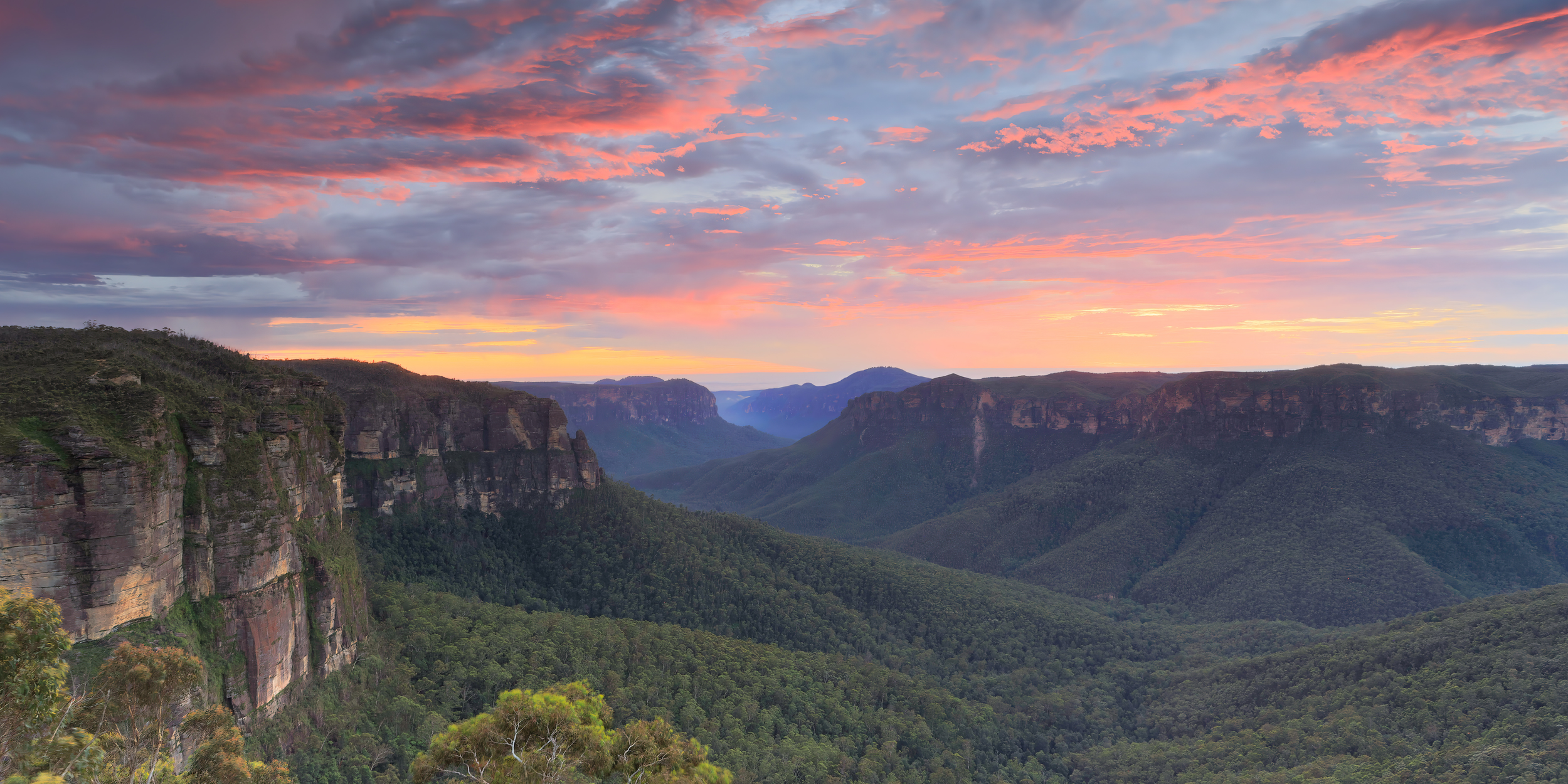 Scenic view of blue and green mountain ranges with cliffs and dense forests at sunset, pink and orange clouds in the sky.
