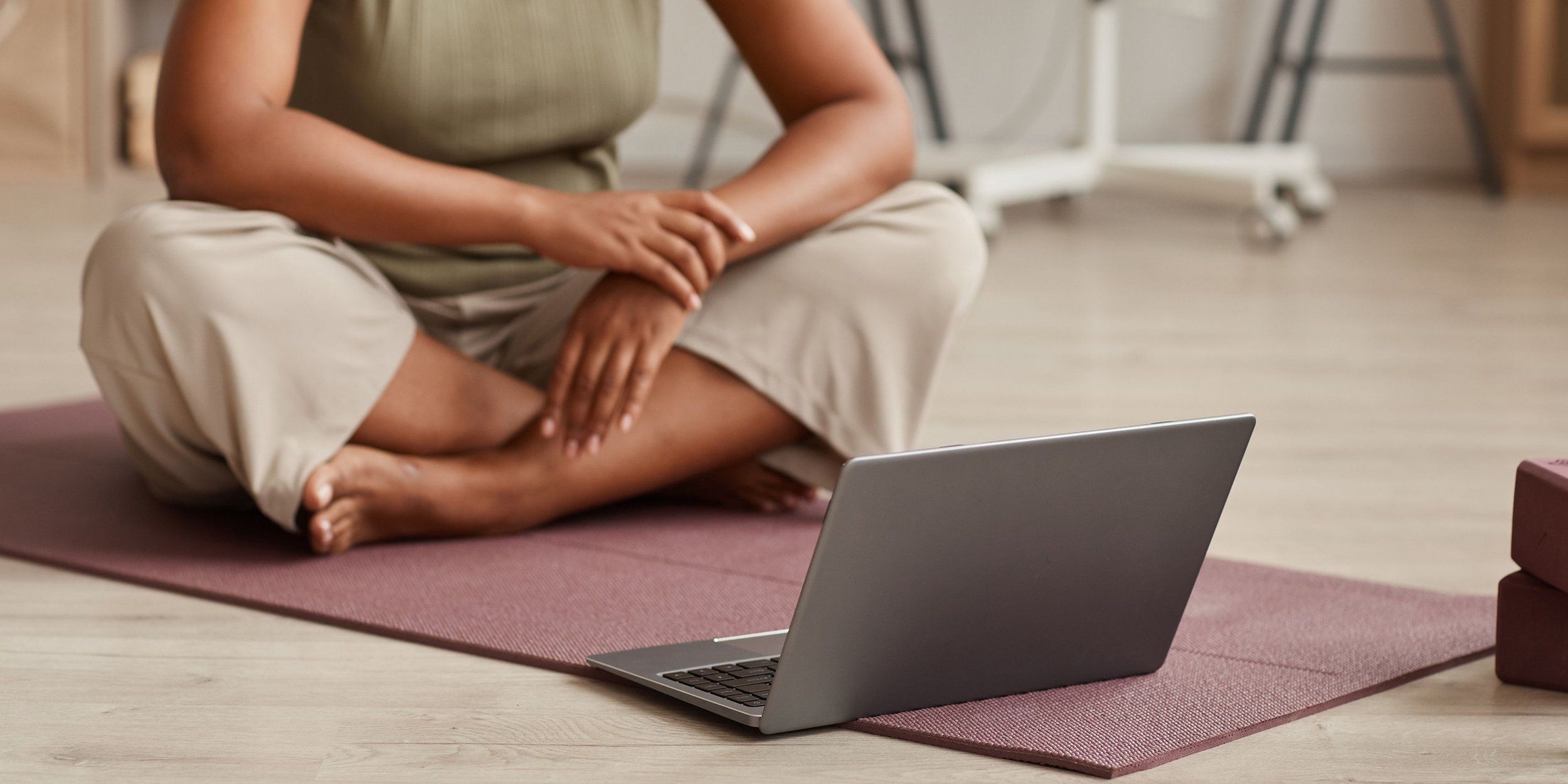 Person sitting cross-legged on yoga mat, using laptop in a room with wooden floor and furniture.