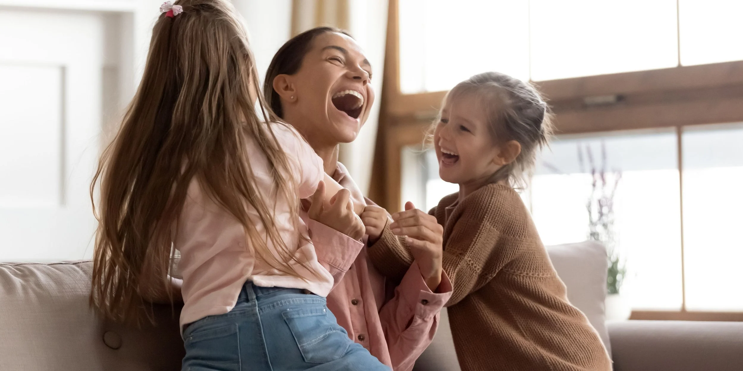 Three females (an adult woman and two young girls) laughing and playing together on a couch in a bright living room.