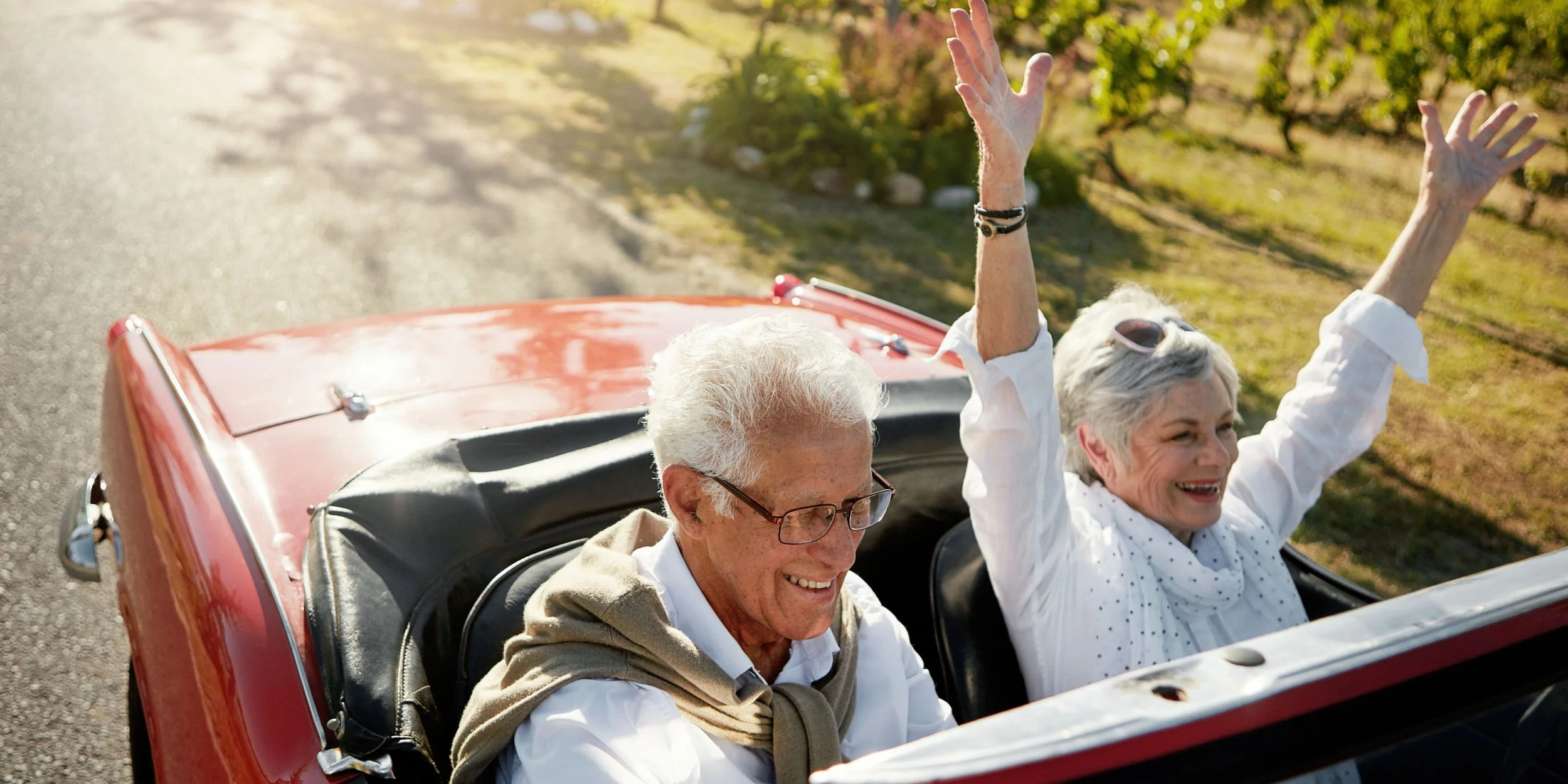 An elderly couple enjoying a drive in a red convertible car with the woman raising her arms in happiness.