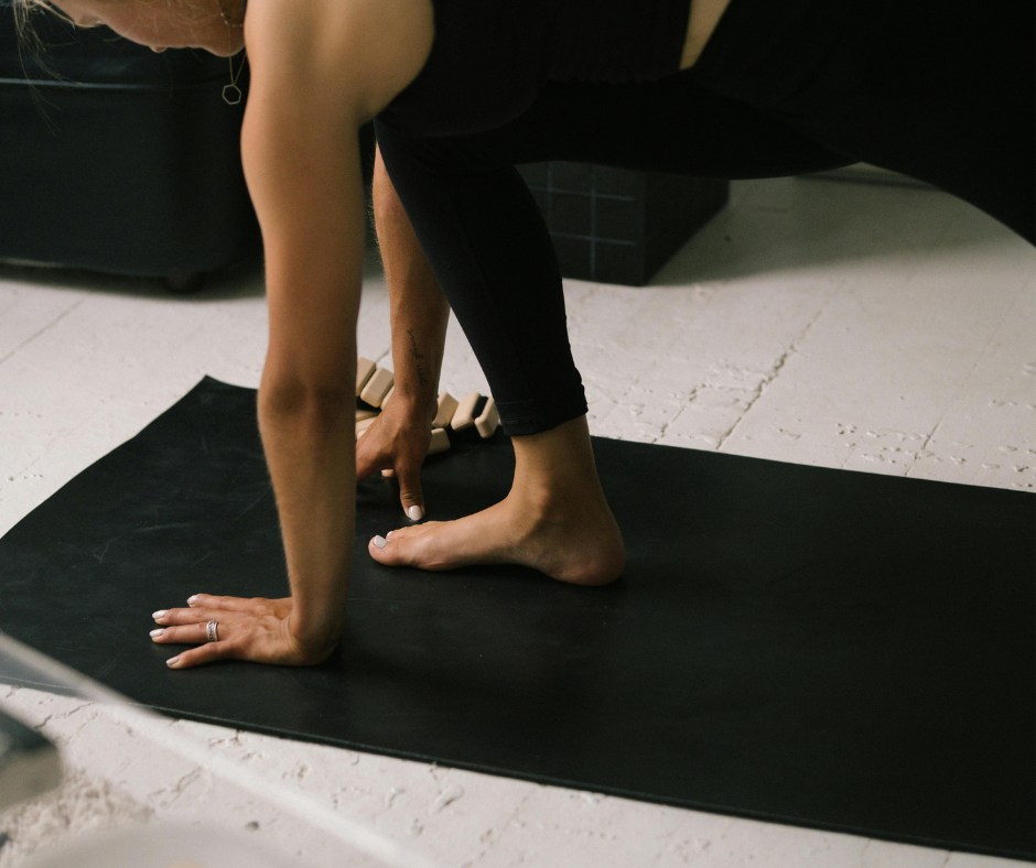 Person practicing yoga or stretching on a black mat indoors, wearing black leggings, with their hands and feet on the floor.