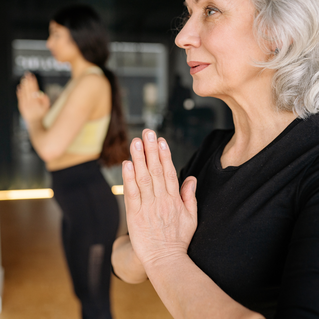 An older woman with gray hair and a black shirt, with her hands pressed together in a prayer position, in a yoga or meditation pose, with another woman with long dark hair and a yellow sports bra practicing yoga or meditation in the background.