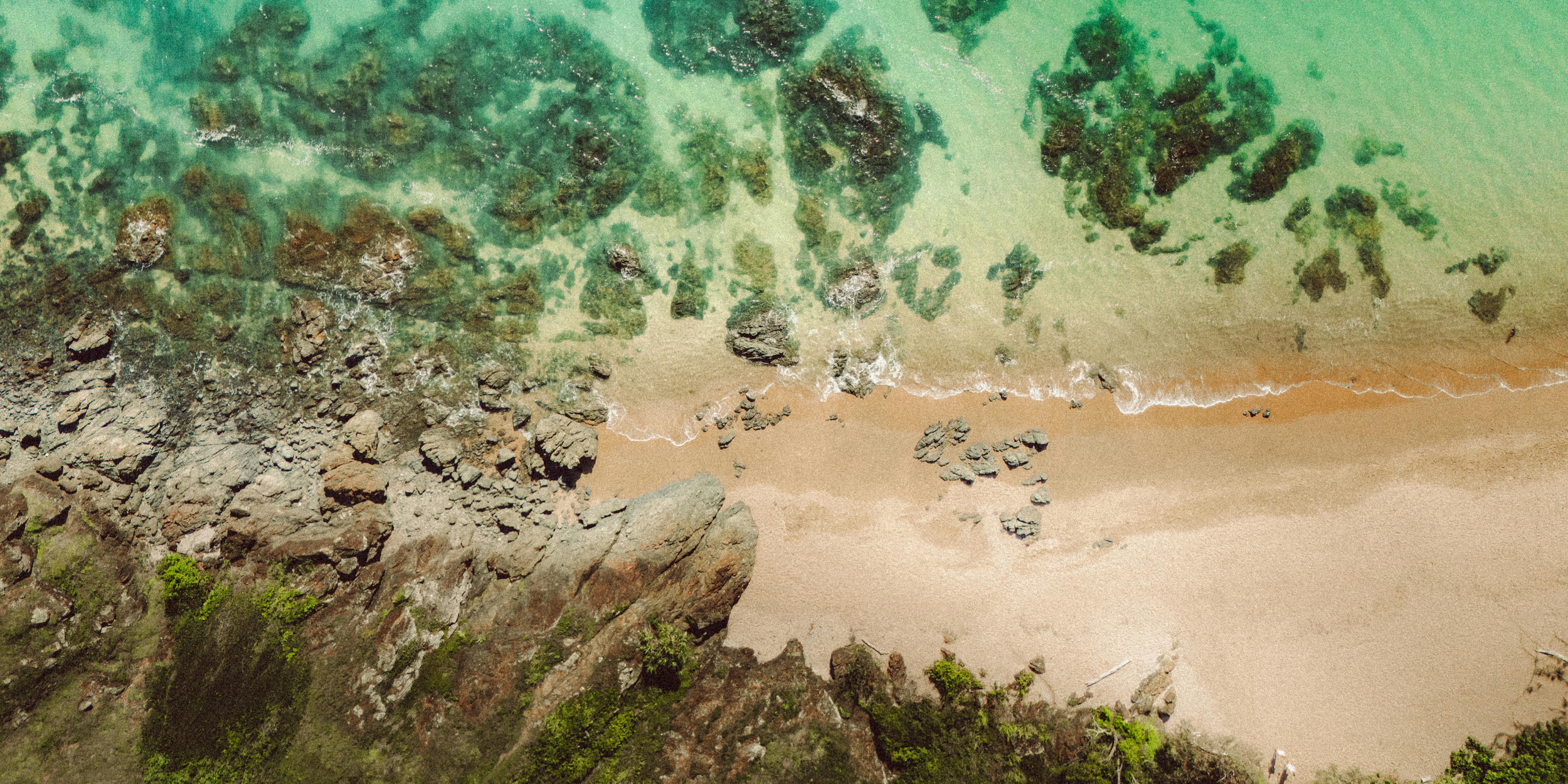 Aerial view of a sandy beach with rocks and green moss, meeting clear green ocean water.