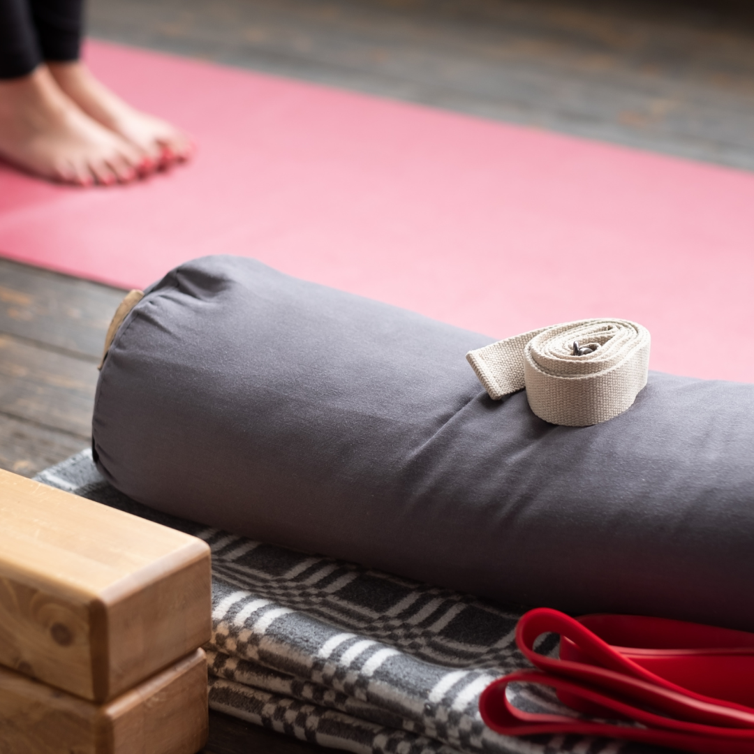 A yoga space with a pink mat, a person's feet visible, and a gray bolster with a rolled white strap on it, placed on a striped blanket on a wooden floor, with red yoga straps nearby.