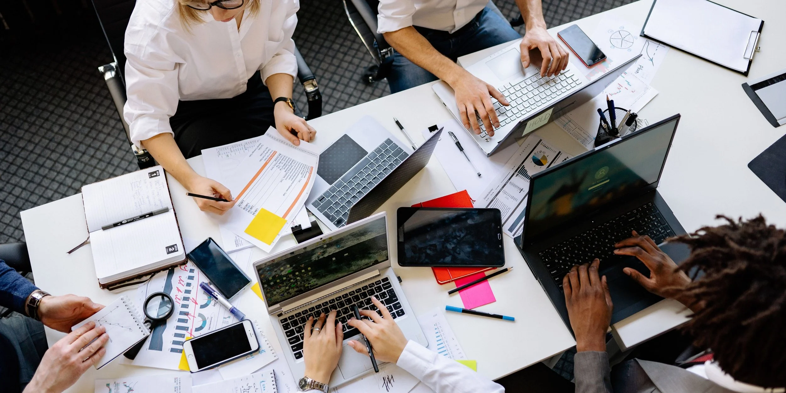 Overview of a busy business meeting with people working on laptops, tablets, and paperwork at a white table.