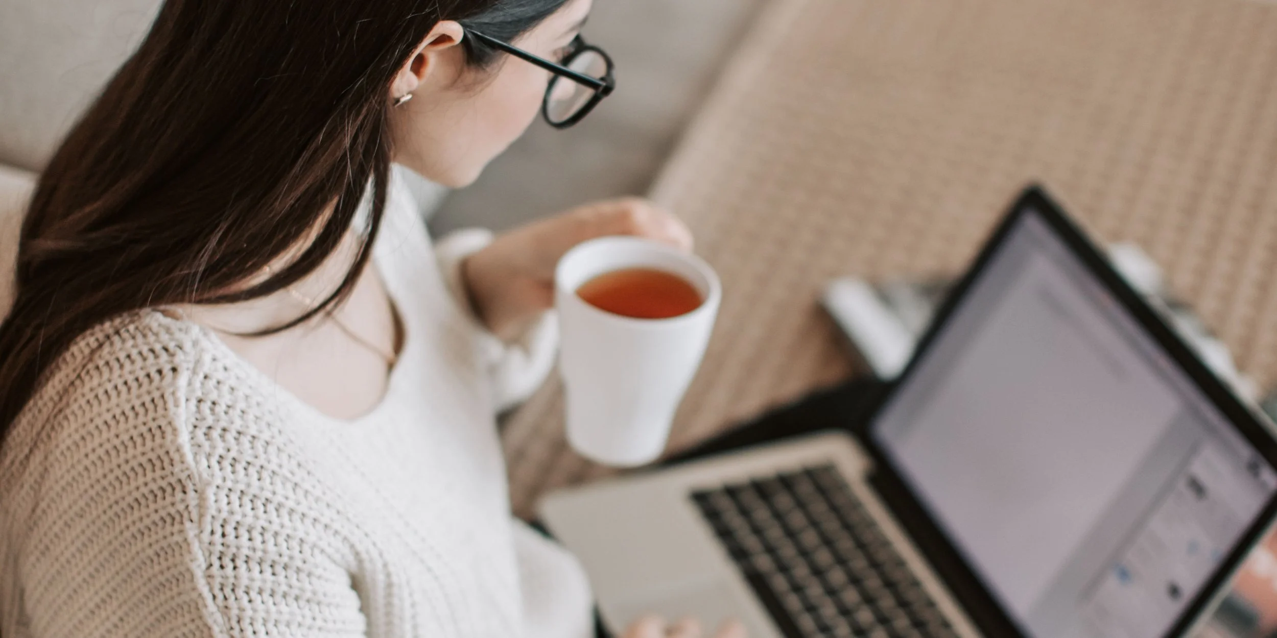 A woman with long brown hair, wearing glasses and a white knitted sweater, sitting on a couch with a cup of tea in her right hand while looking at her laptop.