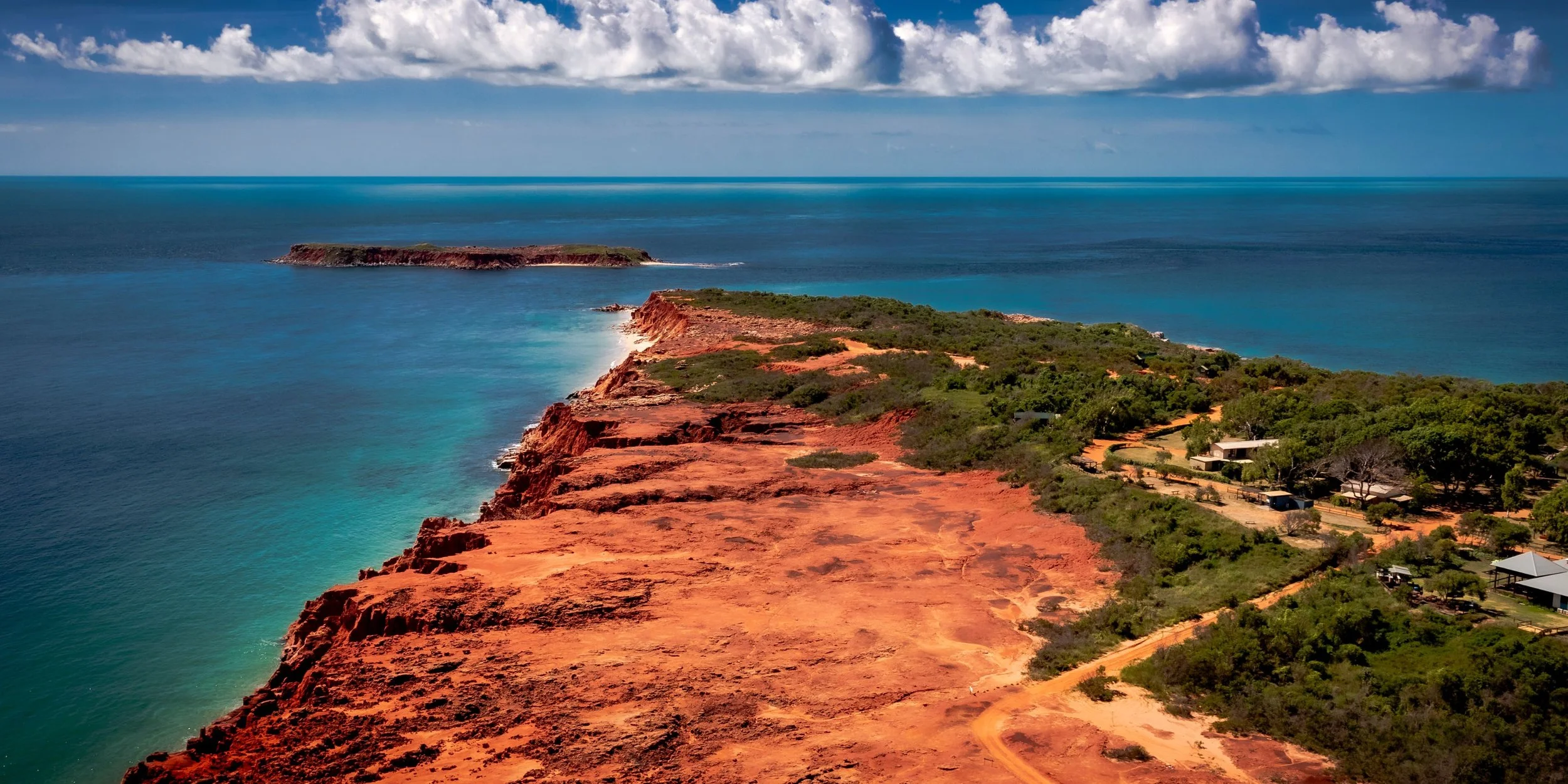 Red rocky coastline with green vegetation and some buildings, overlooking blue ocean under partly cloudy sky.