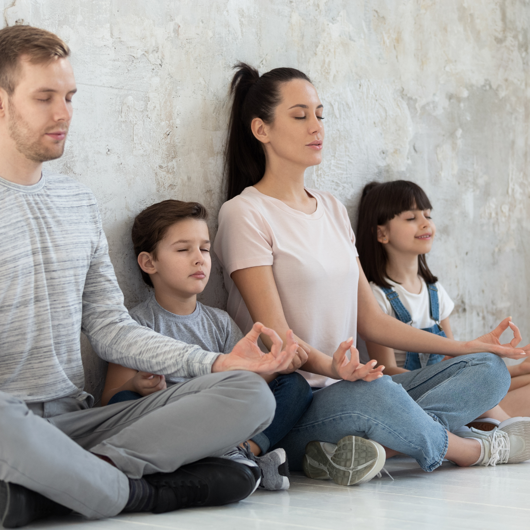 A family of five sitting cross-legged against a textured wall, meditating with eyes closed and hands resting on knees in a serene pose.