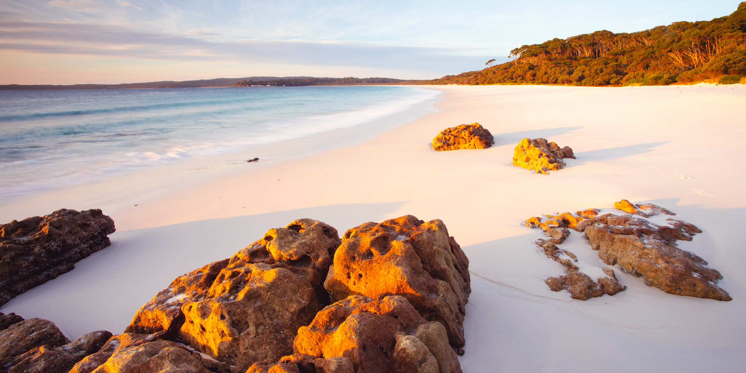 Sunlit rocky beach with white sand, ocean waves, and distant tree-covered coastline at sunset.