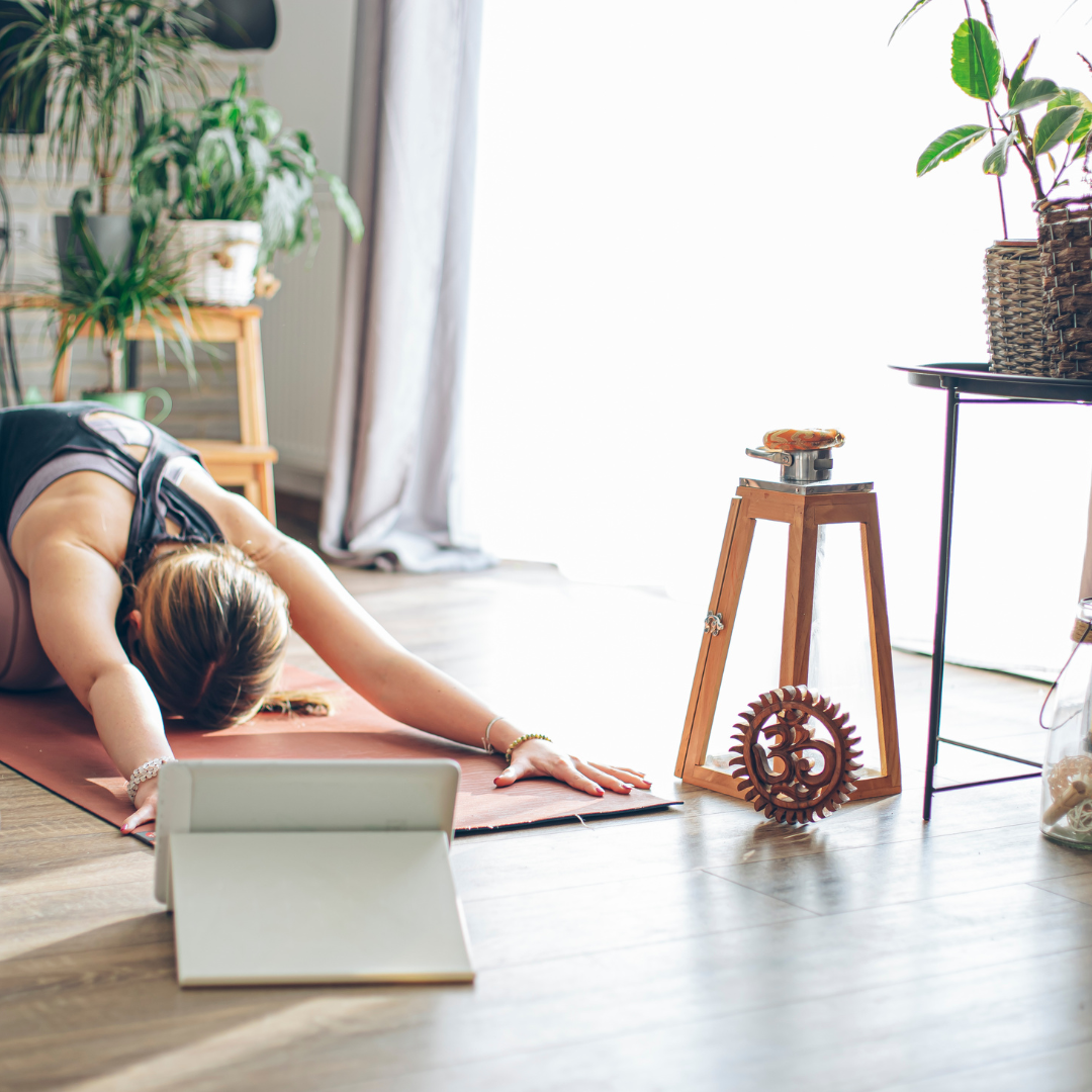 A woman practicing yoga on a mat indoors near a window with natural light, surrounded by houseplants and decorative items.