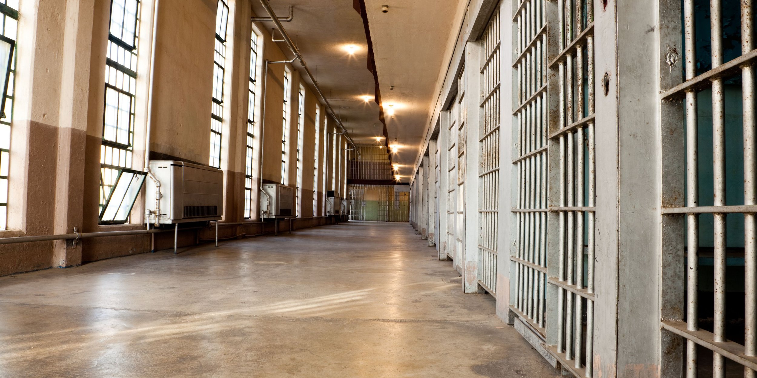 View of a prison corridor with barred prison cells on the right, large windows on the left letting in natural light, and a polished concrete floor.