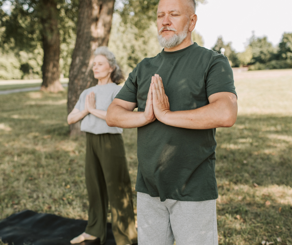 An older man and woman practicing yoga outdoors, standing in a praying position with their eyes closed, in a park or garden setting with trees and grass.
