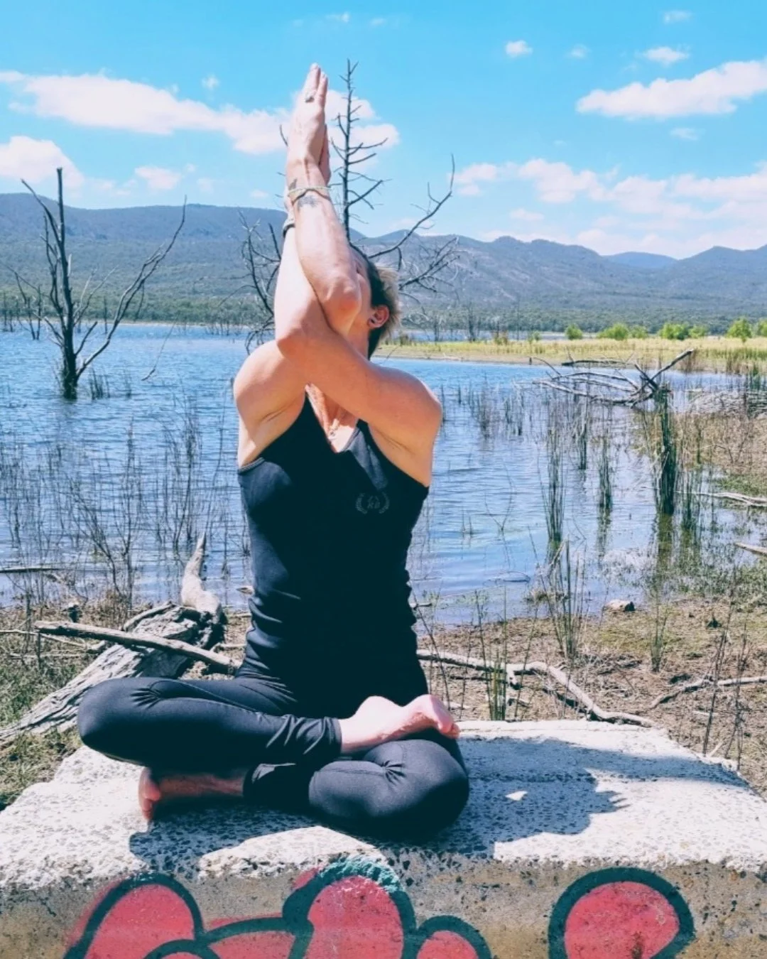 A person practicing yoga outdoors on a concrete ledge by a lake, surrounded by trees and mountains, with a blue sky and scattered clouds.