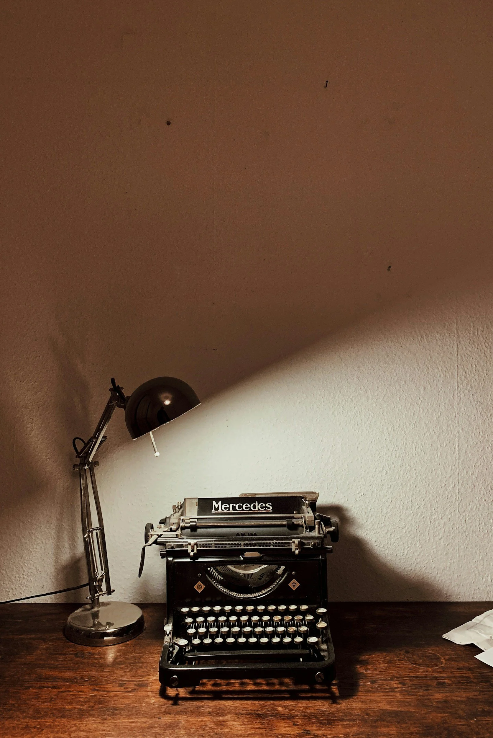 Vintage black typewriter with the word 'Mercedes' on a label, placed on a wooden desk next to a modern silver desk lamp, against a beige wall.