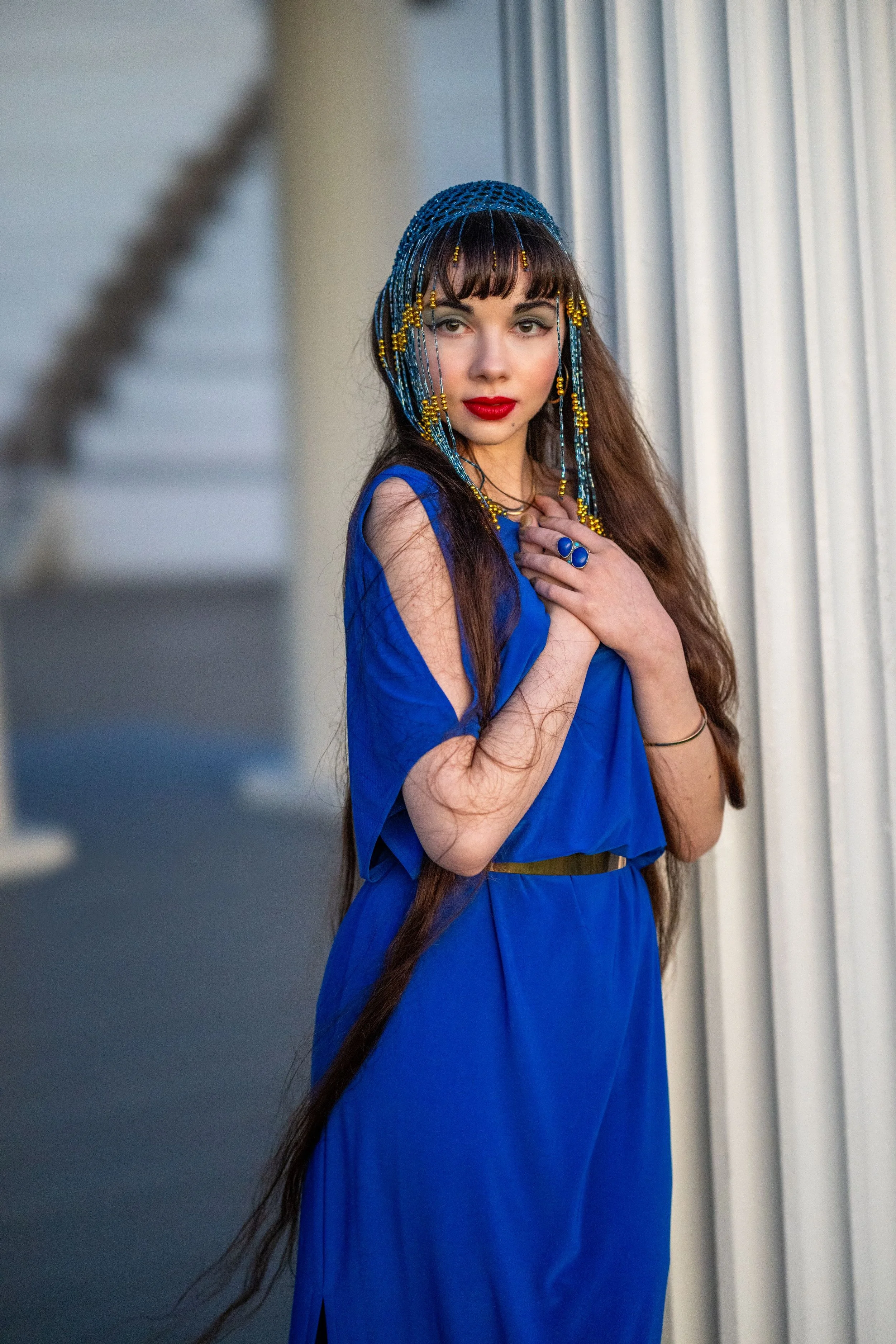 Young woman in blue dress with long dark hair, wearing beaded headpiece and jewelry, standing by white pillars.