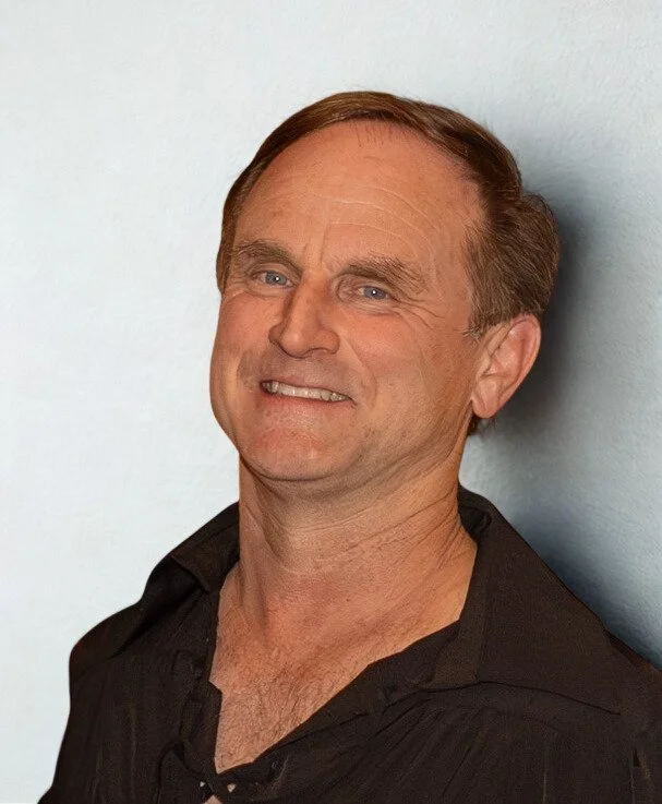 A middle-aged man with light skin and short brown hair smiling, wearing a black shirt, standing against a light blue wall.