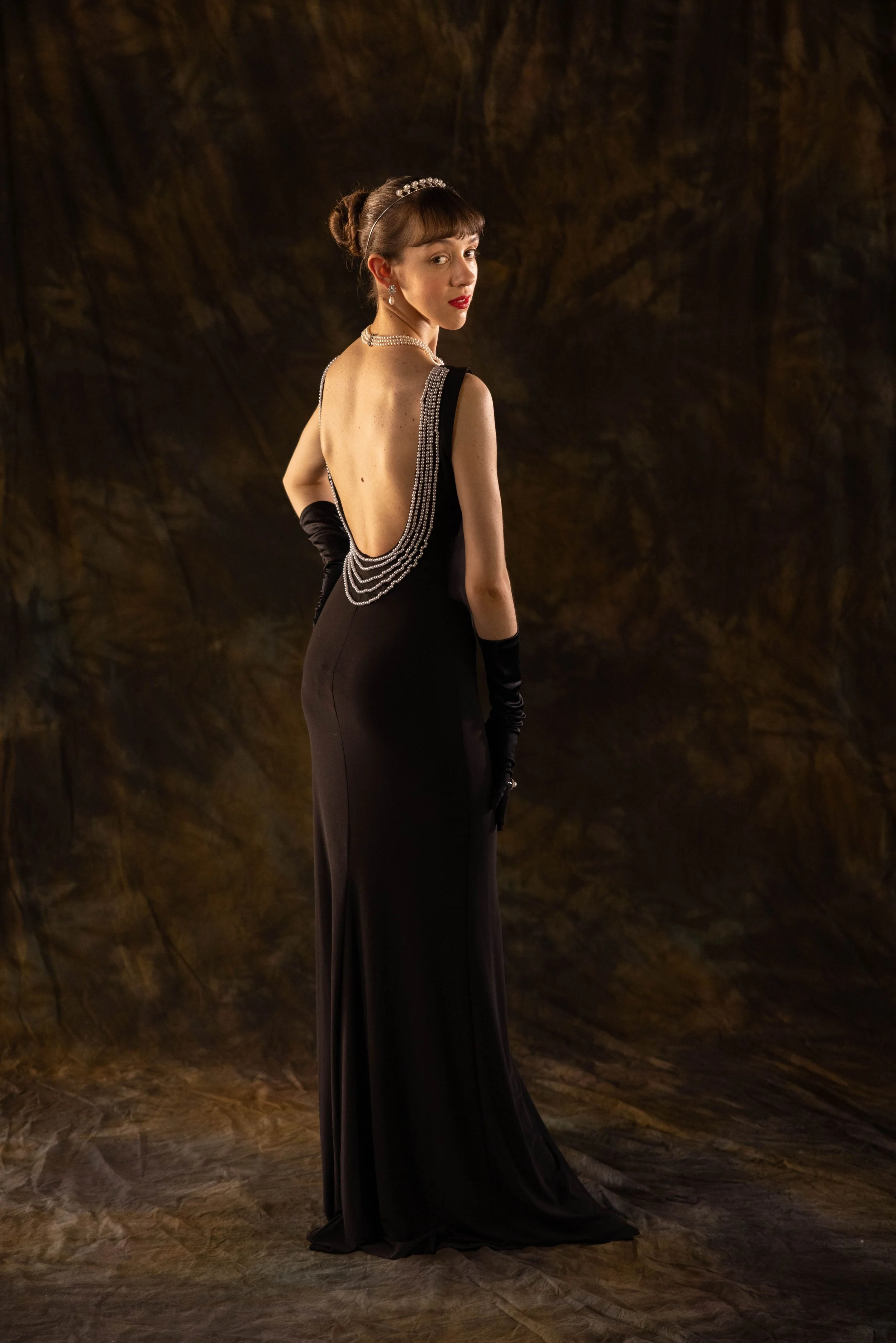 A woman in a black evening gown with a deep open back, wearing long black gloves and pearl jewelry, standing against a dark background.