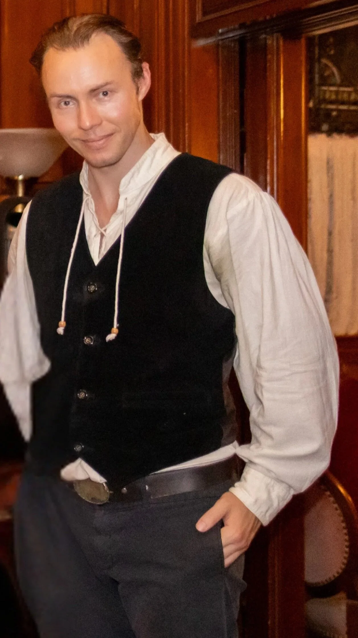 A young man dressed in historical clothing, wearing a white shirt and black vest, standing in a room with wooden decor, smiling at the camera.