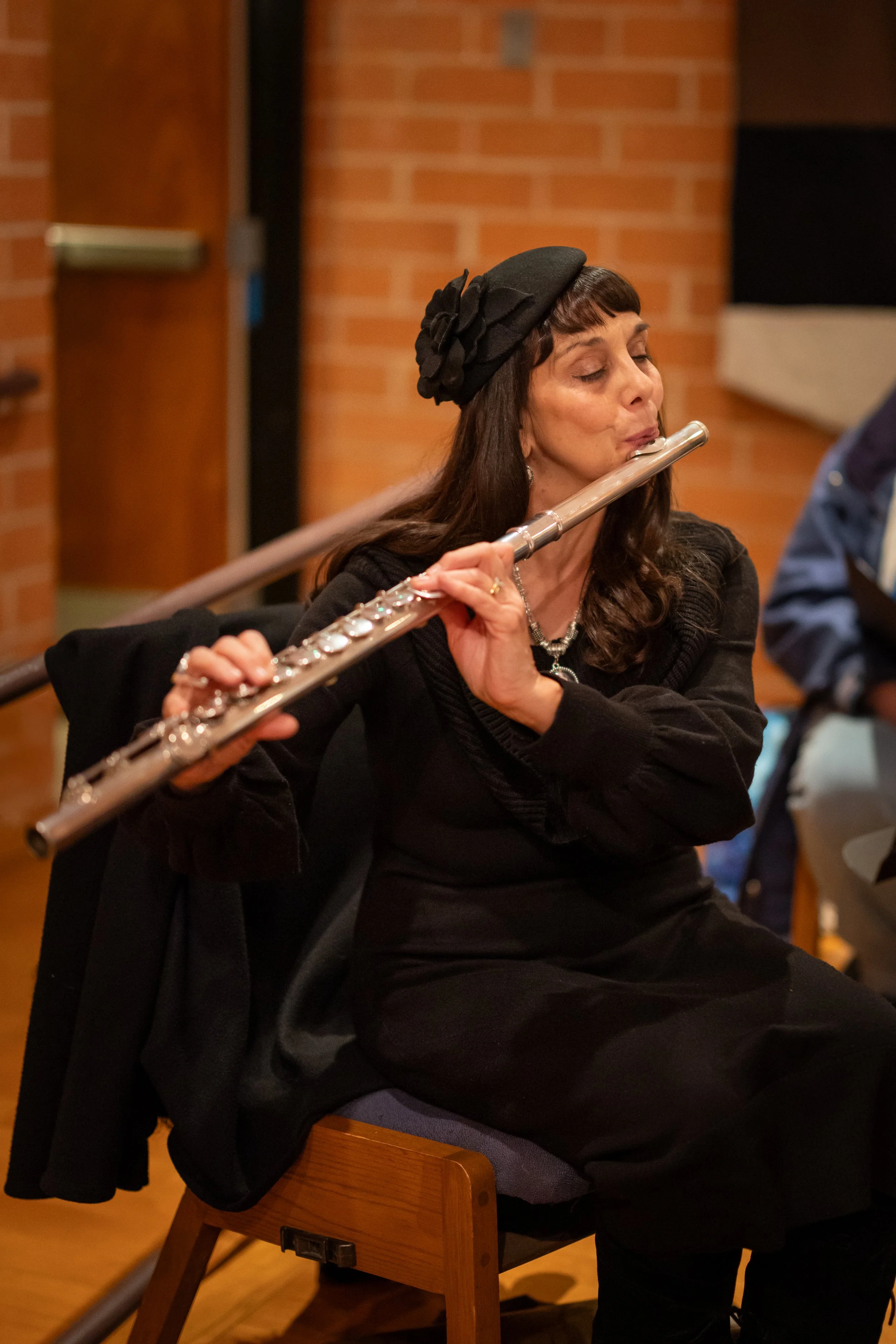Woman wearing a black dress and hat playing a silver flute indoors.
