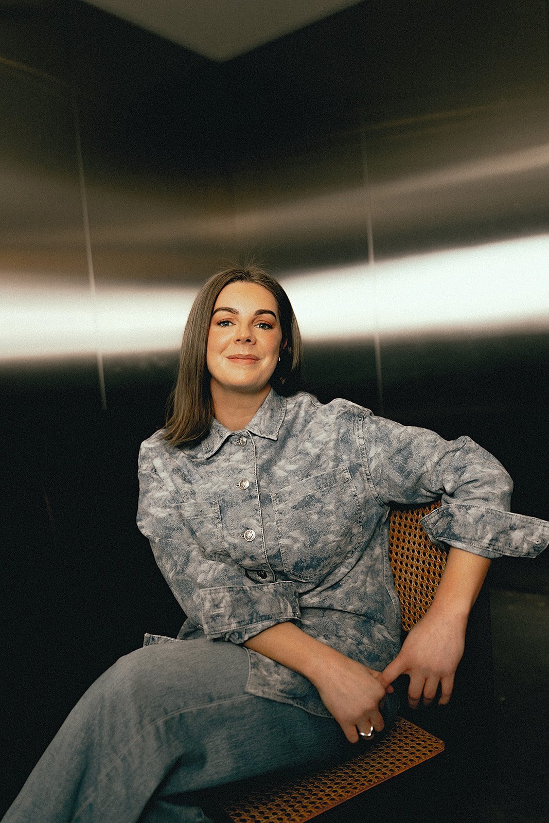 A woman in double denim smiling and sitting on a chair in front of a metal background.