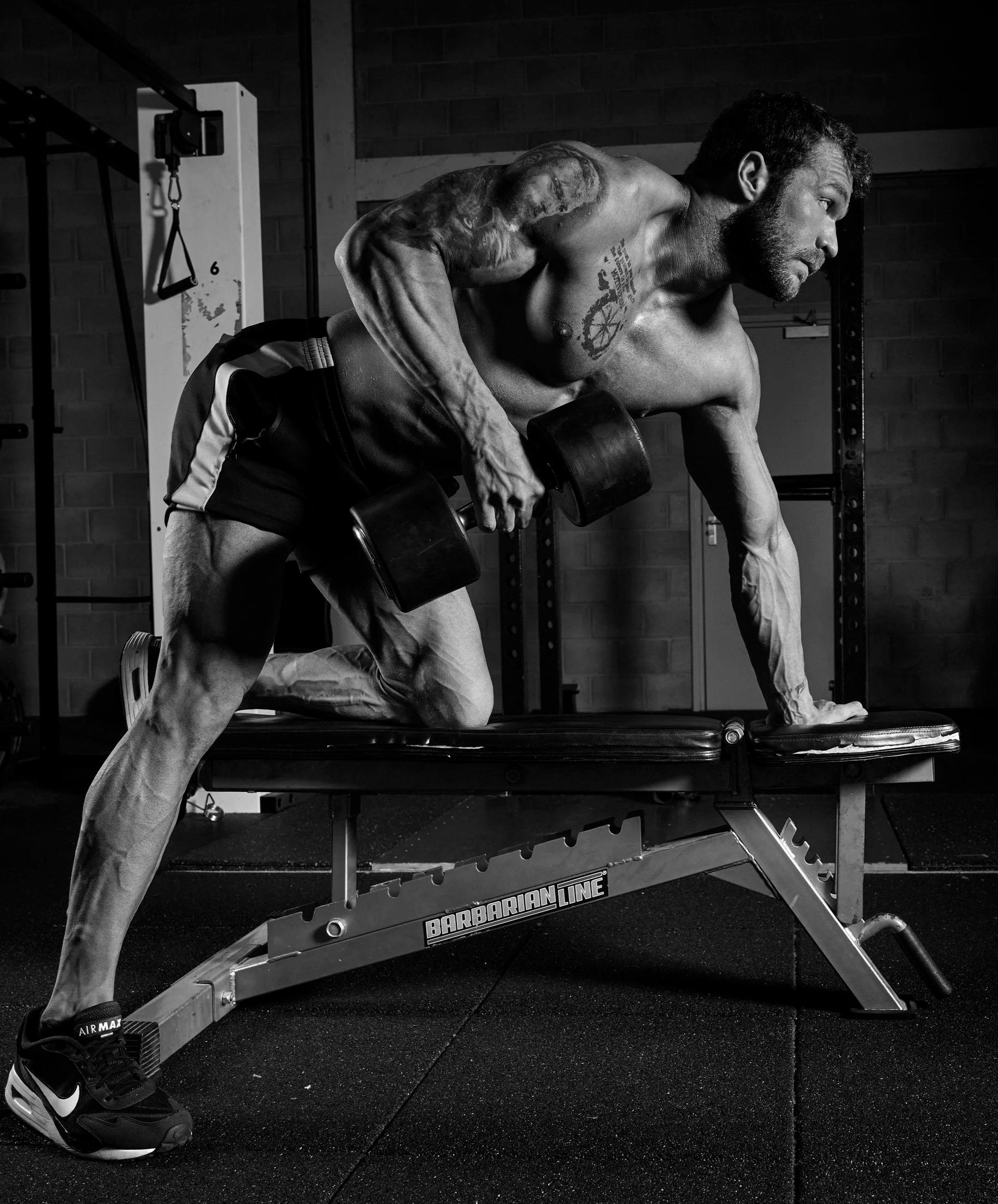 A muscular man with tattoos, shirtless, performing a bent-over row exercise with dumbbells on a bench in a gym.
