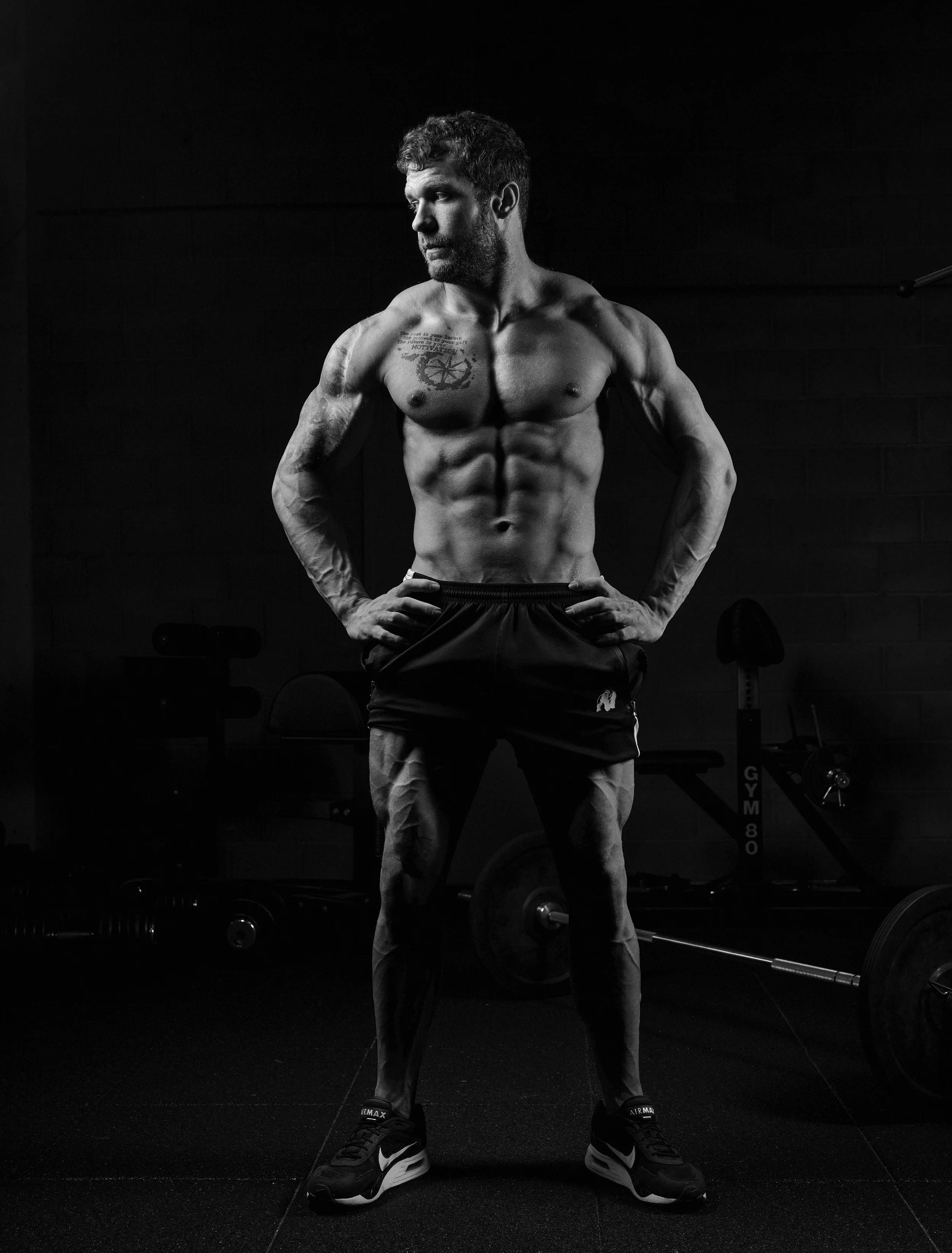 Black and white photo of a muscular man standing with hands on hips in a gym, looking to the side, with barbells and gym equipment in the background.