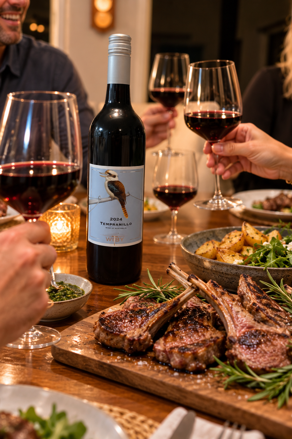 People celebrating with glasses of red wine around a dining table with grilled lamb chops, roasted potatoes, salad, and a bottle of 2024 Tempranillo wine.