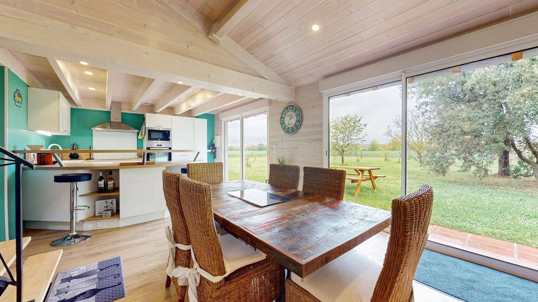 Salle à manger avec table en bois et chaises en rotin, vue vers une cuisine ouverte avec plan de travail blanc, portes-fenêtres donnant sur un jardin avec arbres.