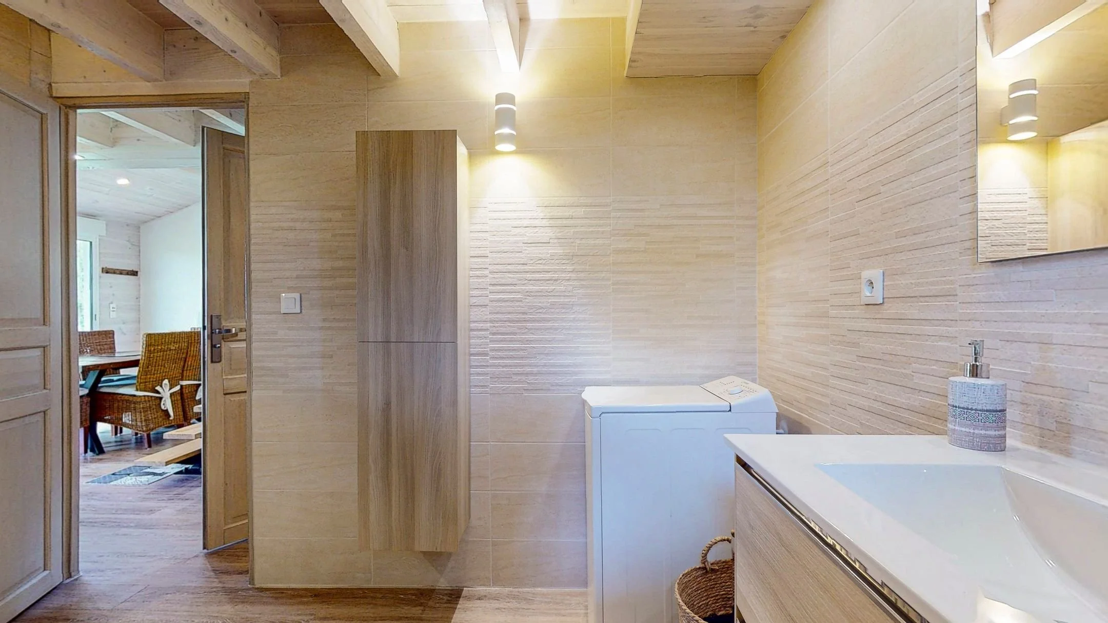 Salle de bain avec lavabo, miroir, sèche-linge blanc, meubles en bois clair, murs en carreaux beige, porte ouverte sur une salle à manger avec chaises en osier et table en bois.