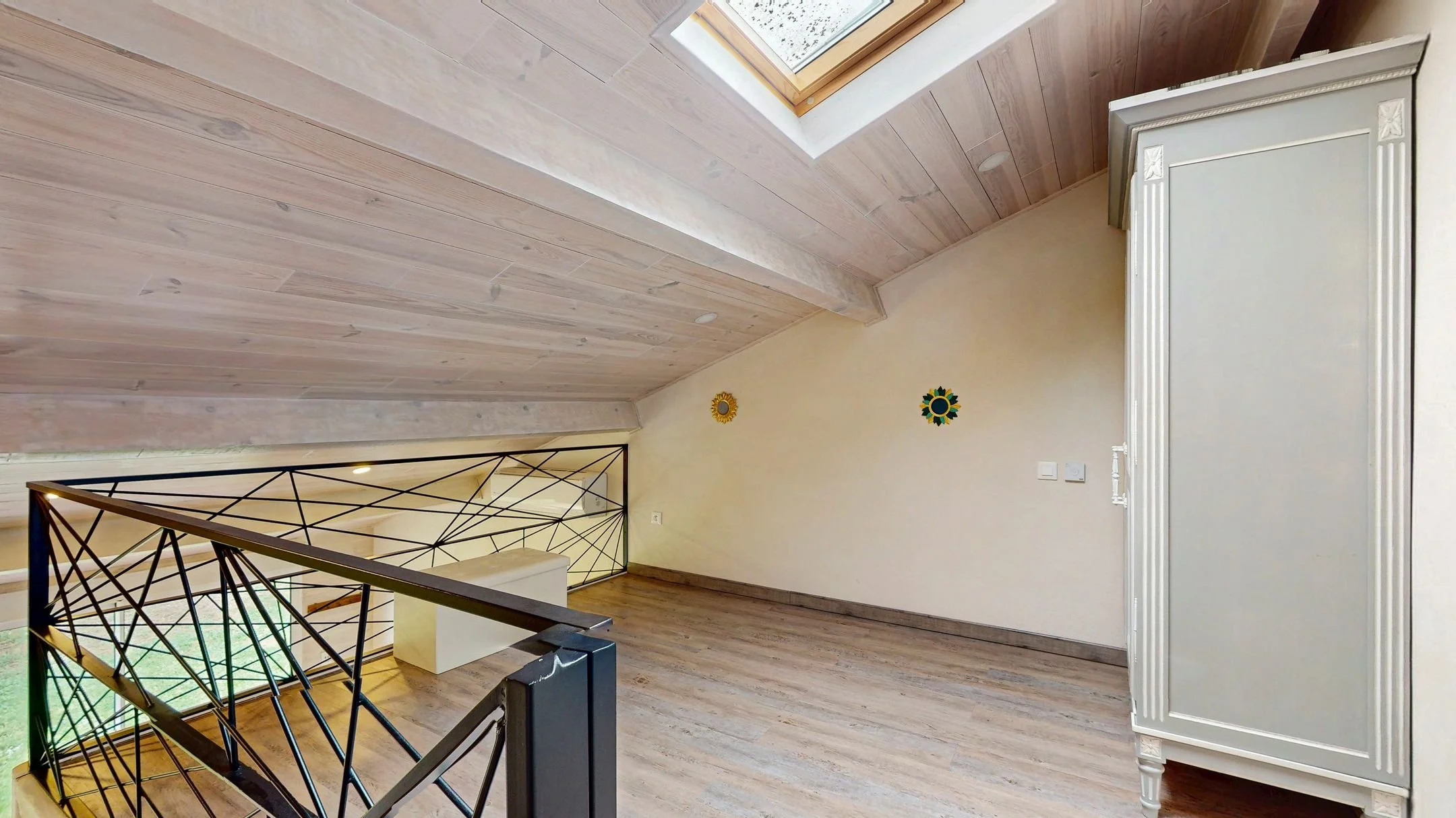Espace de mezzanine avec plafond en bois, un mur beige décoré de deux petits dessins floraux, un grand meuble blanc avec détails sculptés, et un toit en verre permettant la lumière naturelle.