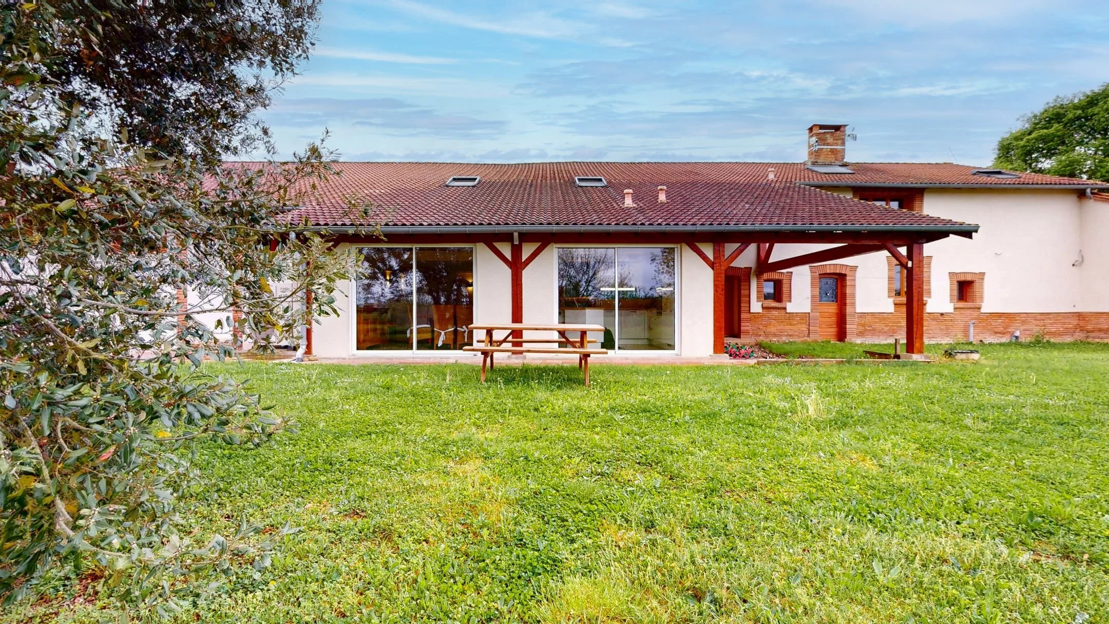 Maison moderne avec terrasse en bois, grandes fenêtres et toit en tuiles rouges, entourée d'un grand espace vert