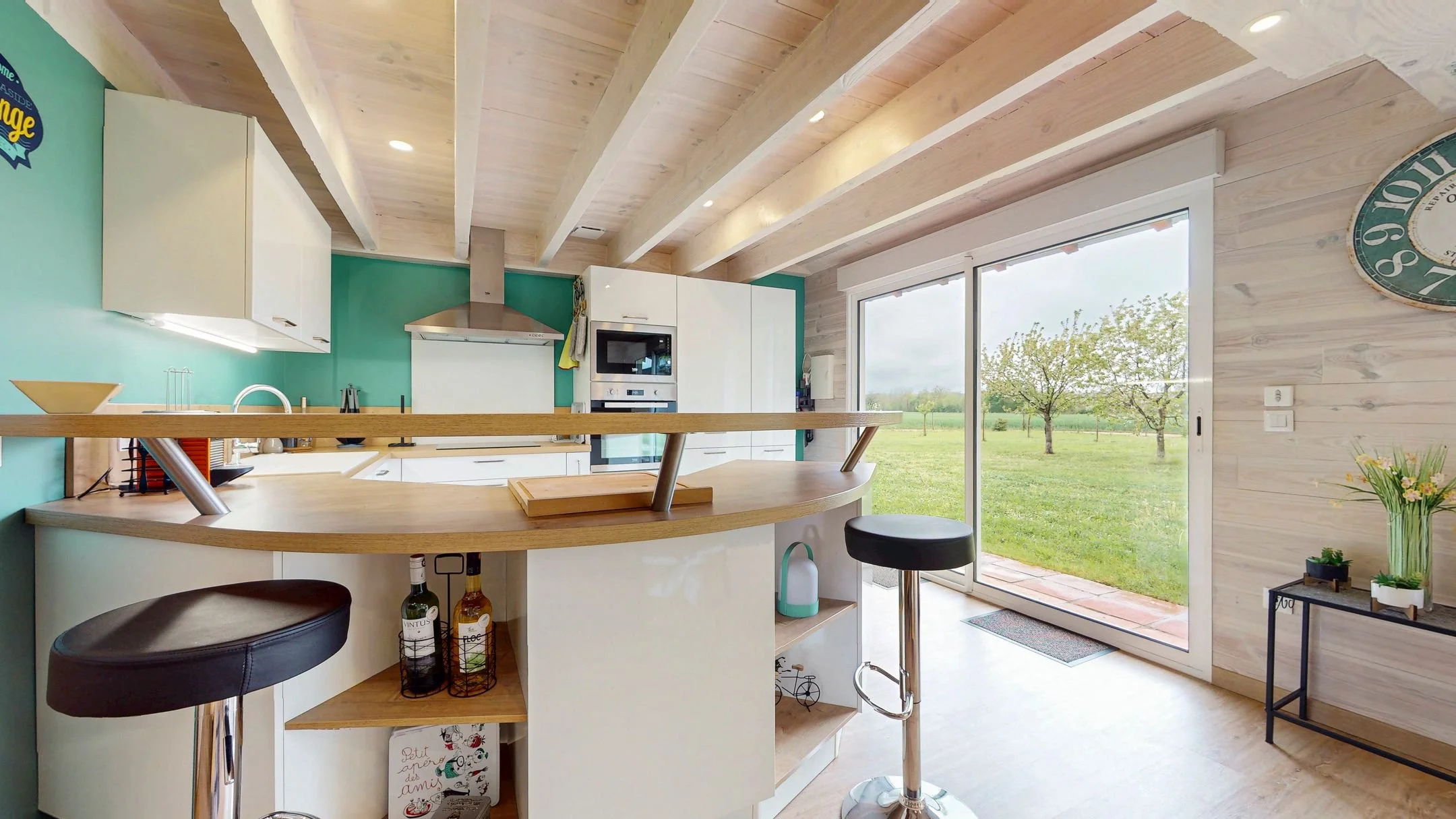 Cuisine moderne avec grande baie vitrée donnant sur un jardin, plans en bois clair, îlot central avec tabourets, armoires blanches, et un horaire mural vert.