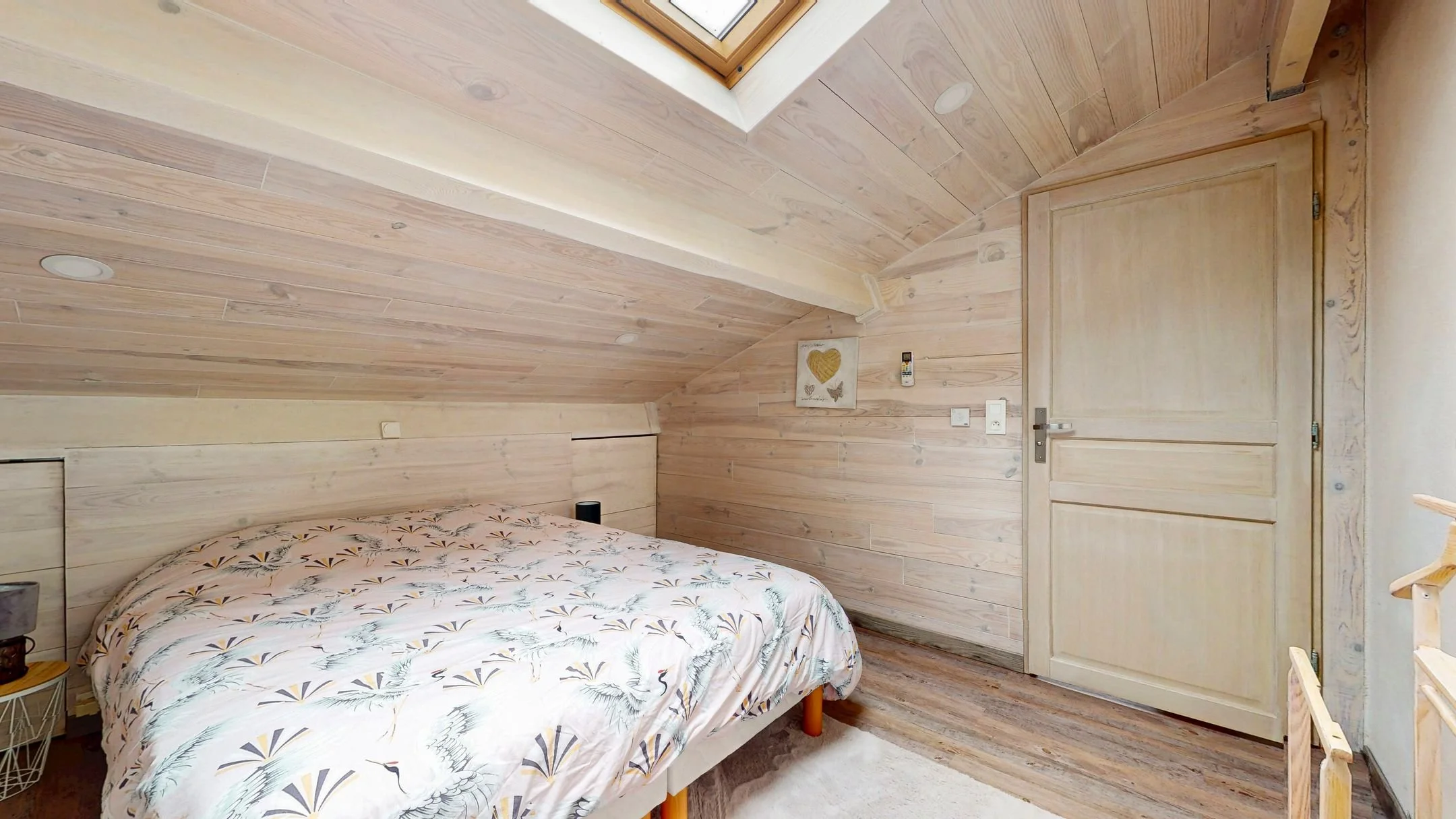 Chambre avec mur en bois clair, lit avec drap à motifs de hérons et feuillages, porte en bois clair, petite décoration murale en forme de cœur, télécommande, et quelques meubles