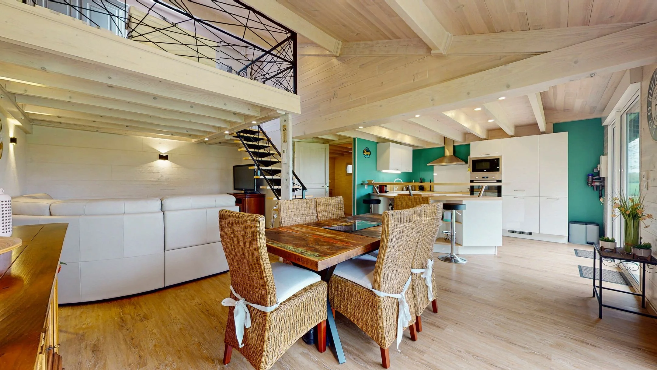 Intérieur spacieux d'une maison avec mezzanine, comprenant une cuisine blanche moderne, une salle à manger avec une table en bois et des chaises en osier, un canapé blanc, un sol en bois clair et des murs en bois clair.