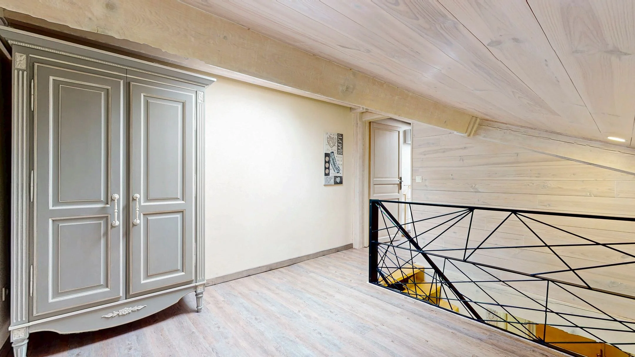 Une mezzanine avec un placard ancien peint en gris, un mur blanc avec une peinture moderne, un panneau de porte et un garde-corps en métal noir avec un design géométrique. Le sol est en bois clair.