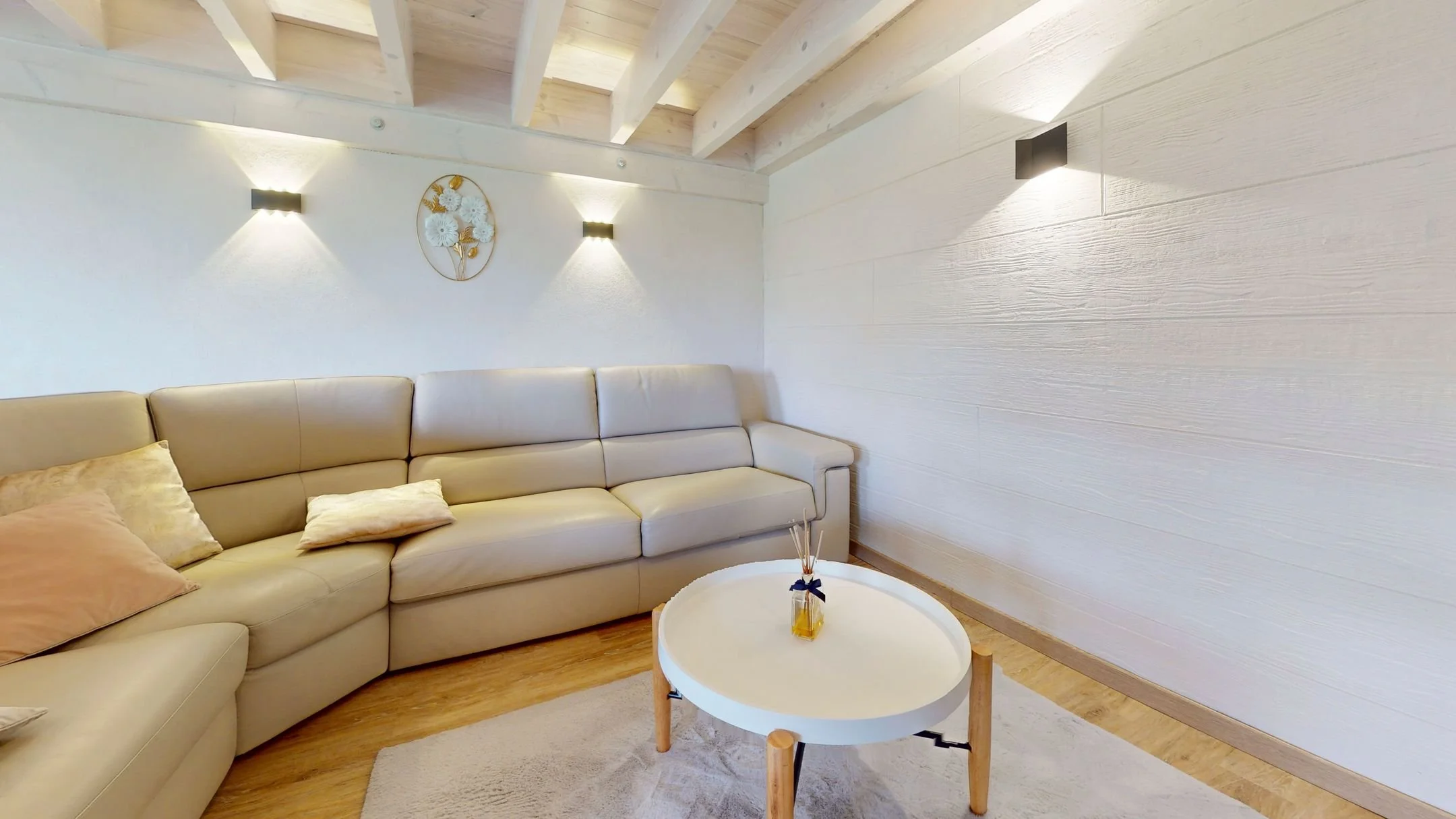 Salon avec canapé beige en forme de L, petits coussins, table basse ronde blanche avec diffuseur parfumé, murs blancs, éclairage mural moderne, plafond en bois clair avec poutres apparentes.