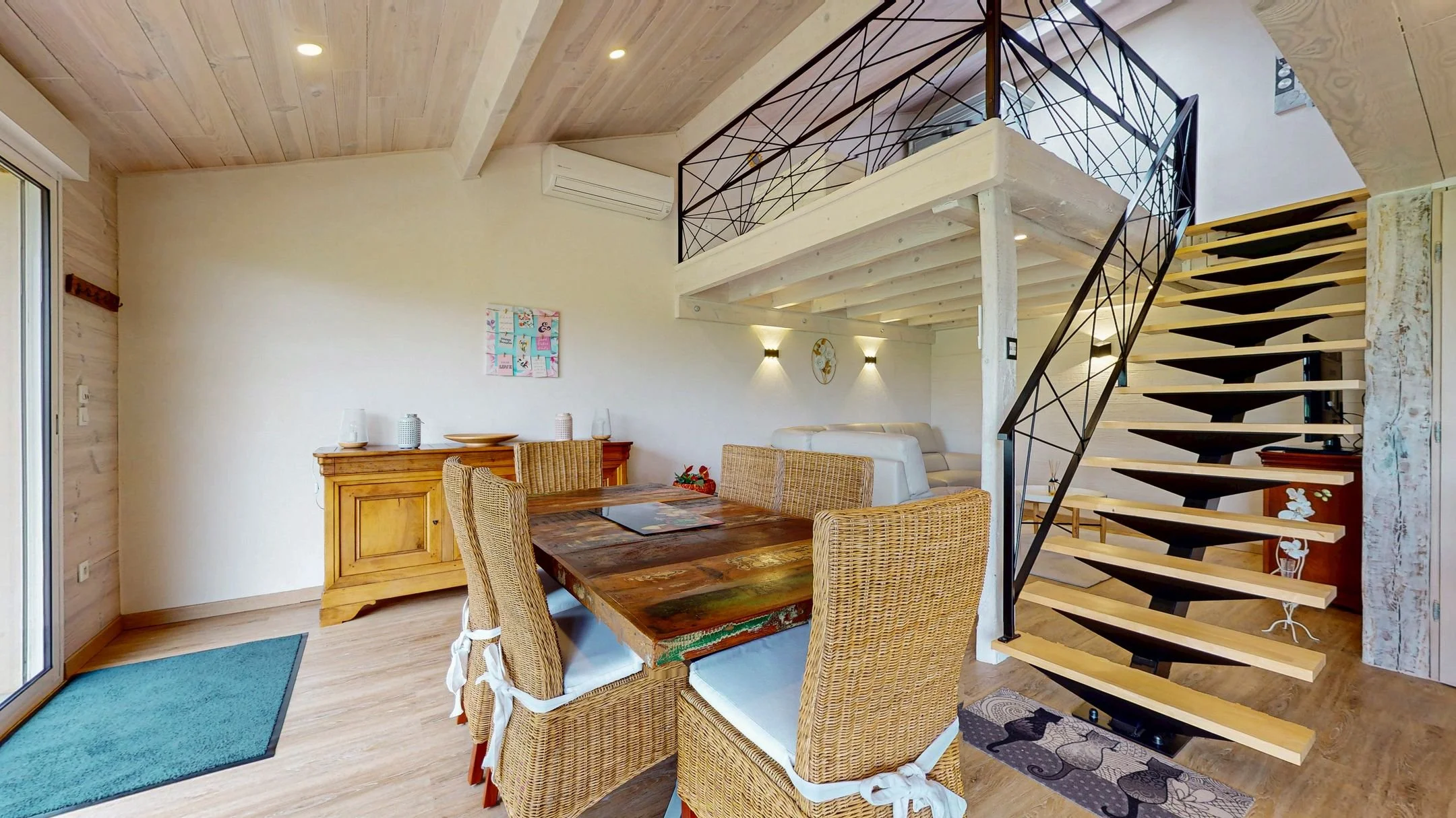 Intérieur d'un salon avec une table en bois, des chaises en rotin et un escalier design en bois et métal.