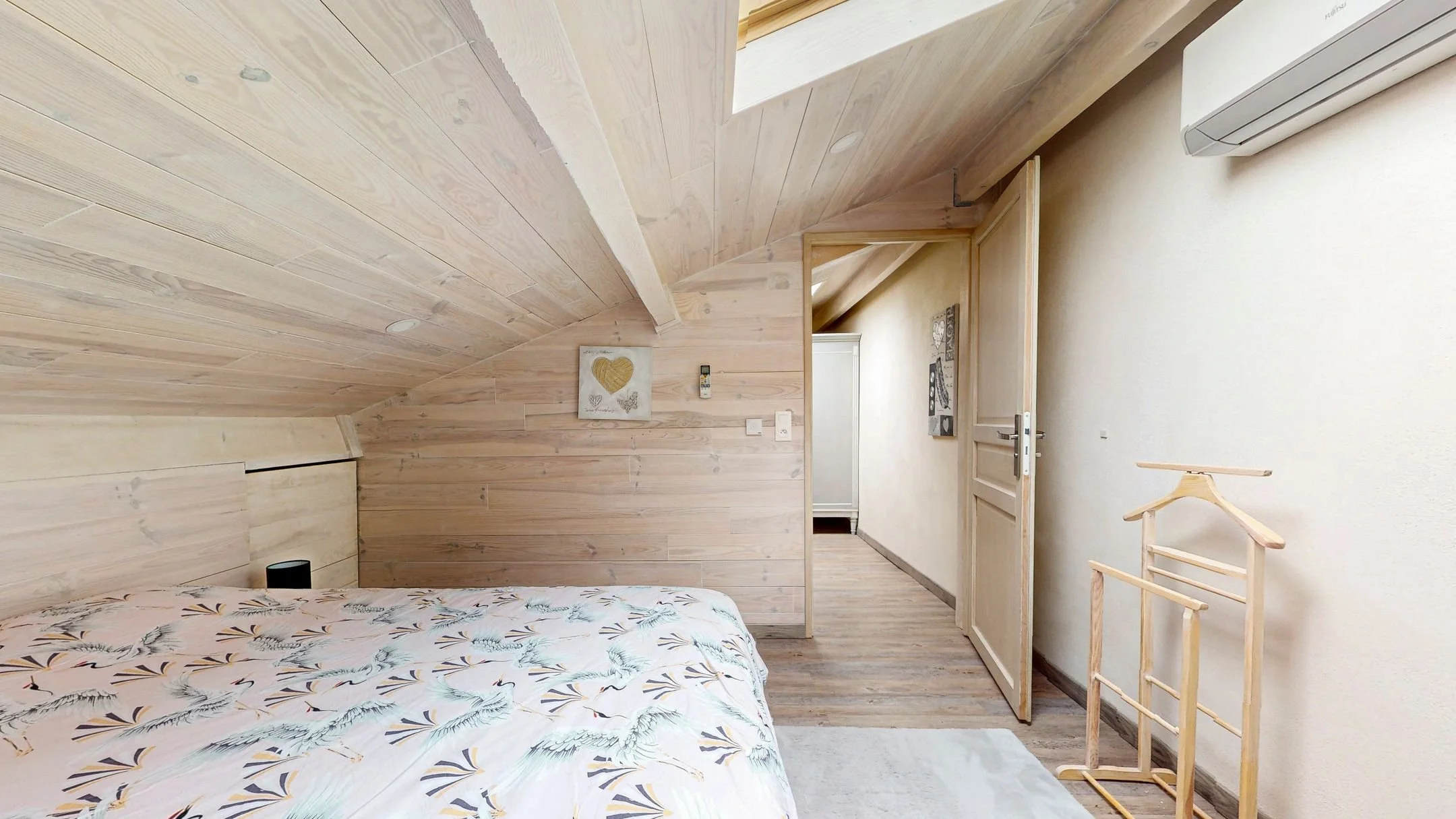 Chambre avec plafond en bois, lit avec drap léger imprimé, porte ouverte menant à un couloir avec décorations murales, ventilateur de plafond blanc, climatiseur, porte-serviettes en bois vide.