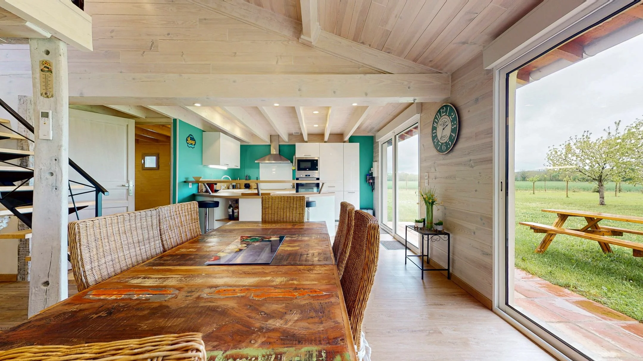 Intérieur d'une maison avec grande table en bois, cuisine ouverte avec armoires blanches, murs turquoise, fenêtres donnant sur un jardin avec pelouse et arbres.