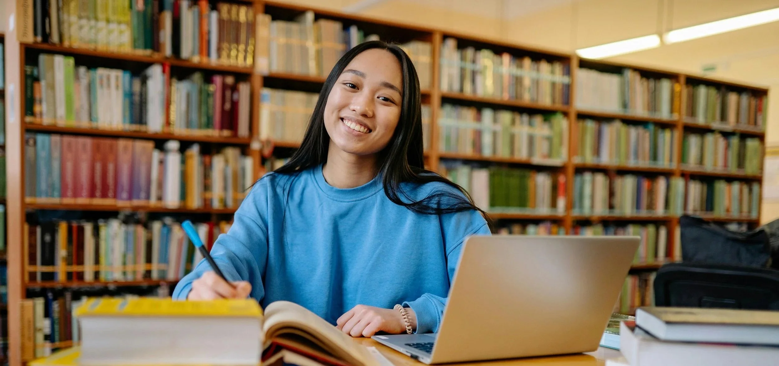 Young woman with long black hair, smiling, sitting at a table in a library, writing with a pen, with a laptop and books around her.
