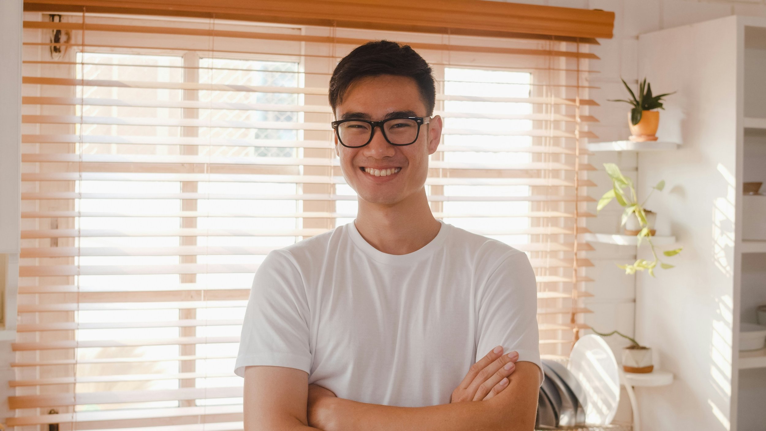 A confident young man wearing glasses smiles at the camera with his arms crossed. He looks approachable and professional, dressed casually in a relaxed setting. This image conveys positivity, self-assurance, and readiness, making it ideal for content
