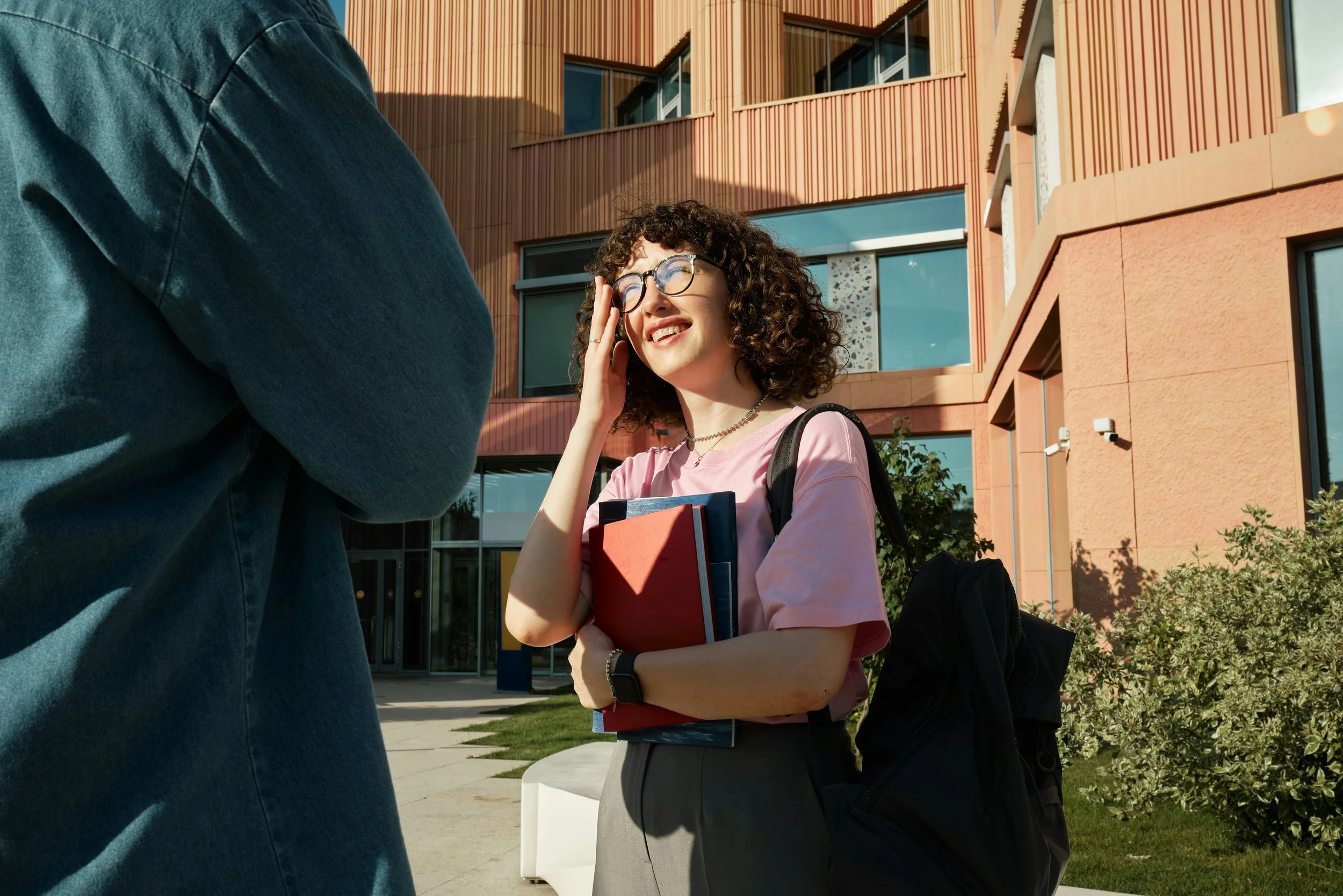 A young woman adjusts her glasses while standing on a sunny college campus. She appears focused and engaged, enjoying the bright outdoor setting. This image is perfect for articles about student life, higher education, university experiences, or camp