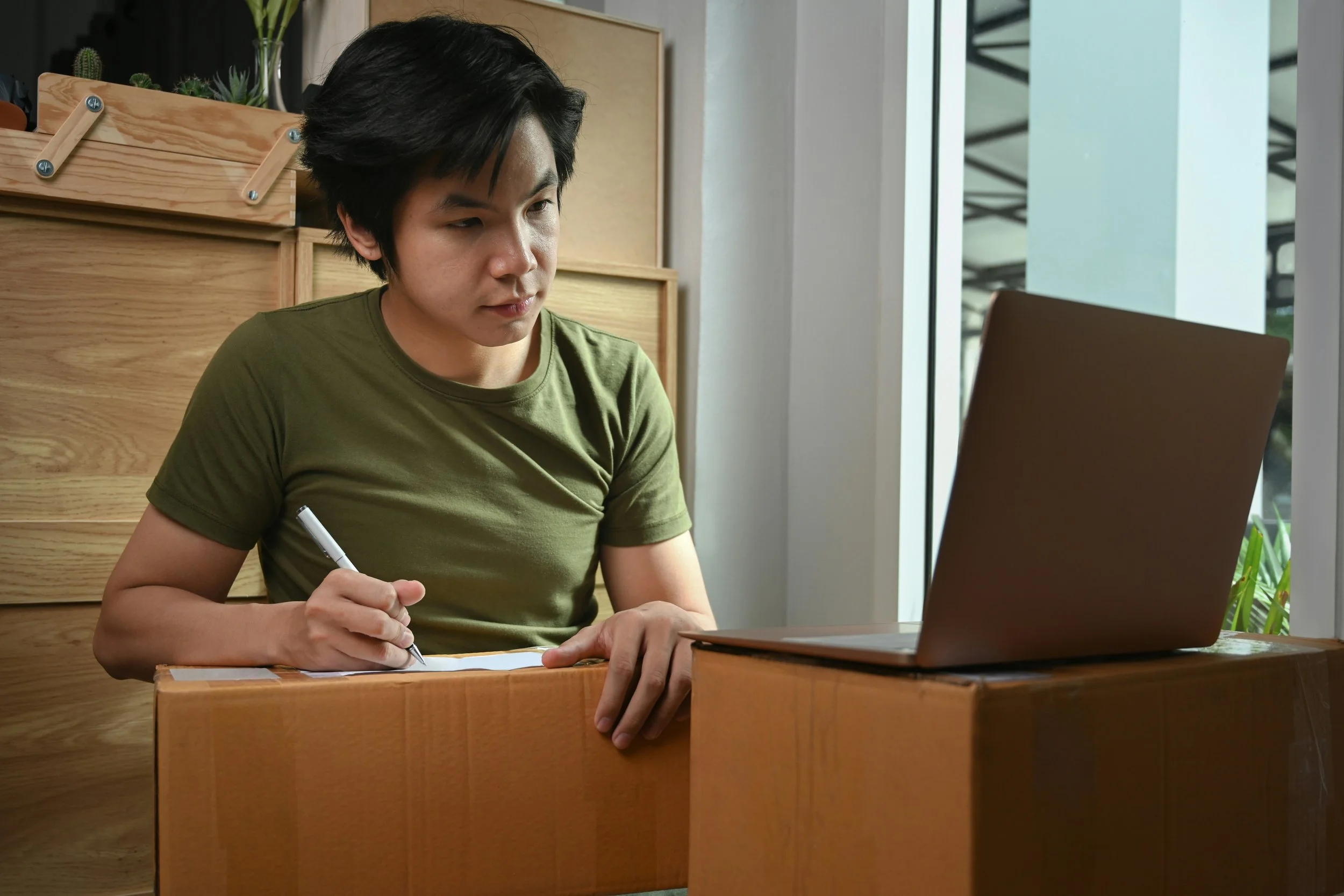 A young man works at a laptop while taking notes, concentrating on his studies. He is seated in a modern workspace, highlighting productivity, learning, and focus. This image works well for content about online learning, study tips, remote work, or p