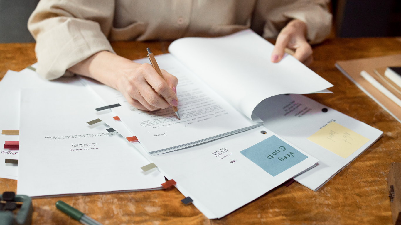 Person sitting at a wooden table, working with documents and papers, holding a pen and turning a page on a binder or notebook.