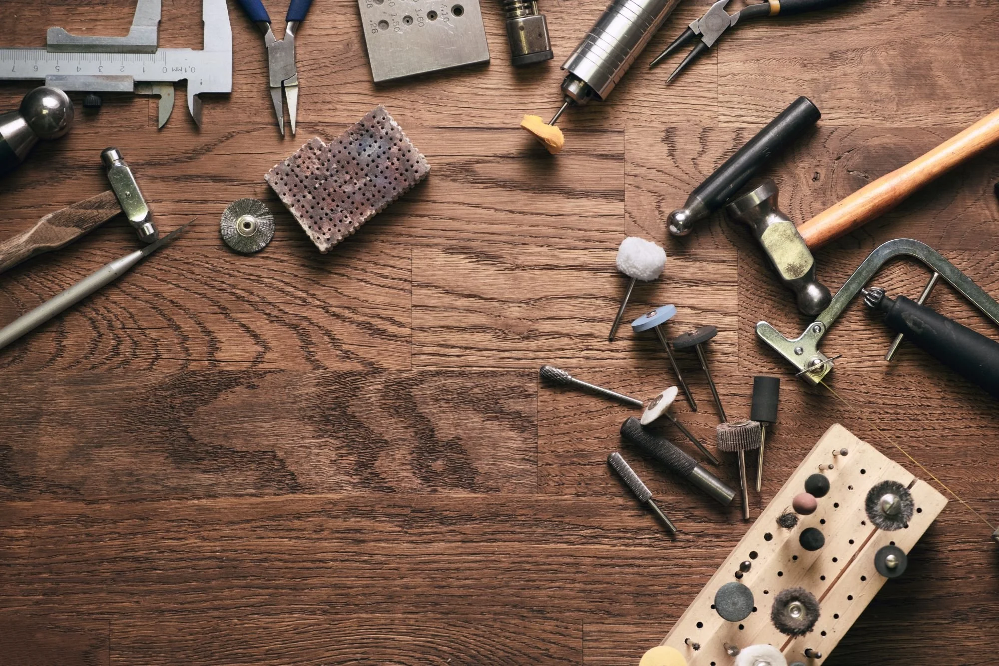 Various jewelry-making tools and supplies on a wooden surface, including files, hammers, polishing wheels, a pegboard with different polishing attachments, and a small workbench with assorted small parts.